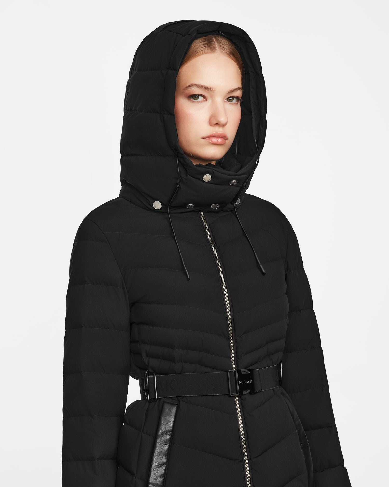 A woman stands slightly to the side against a plain white background, wearing the Rudsak MALAYA M - L123554 Black down puffer jacket with herringbone quilting, hood, zipper front, and belted waist.