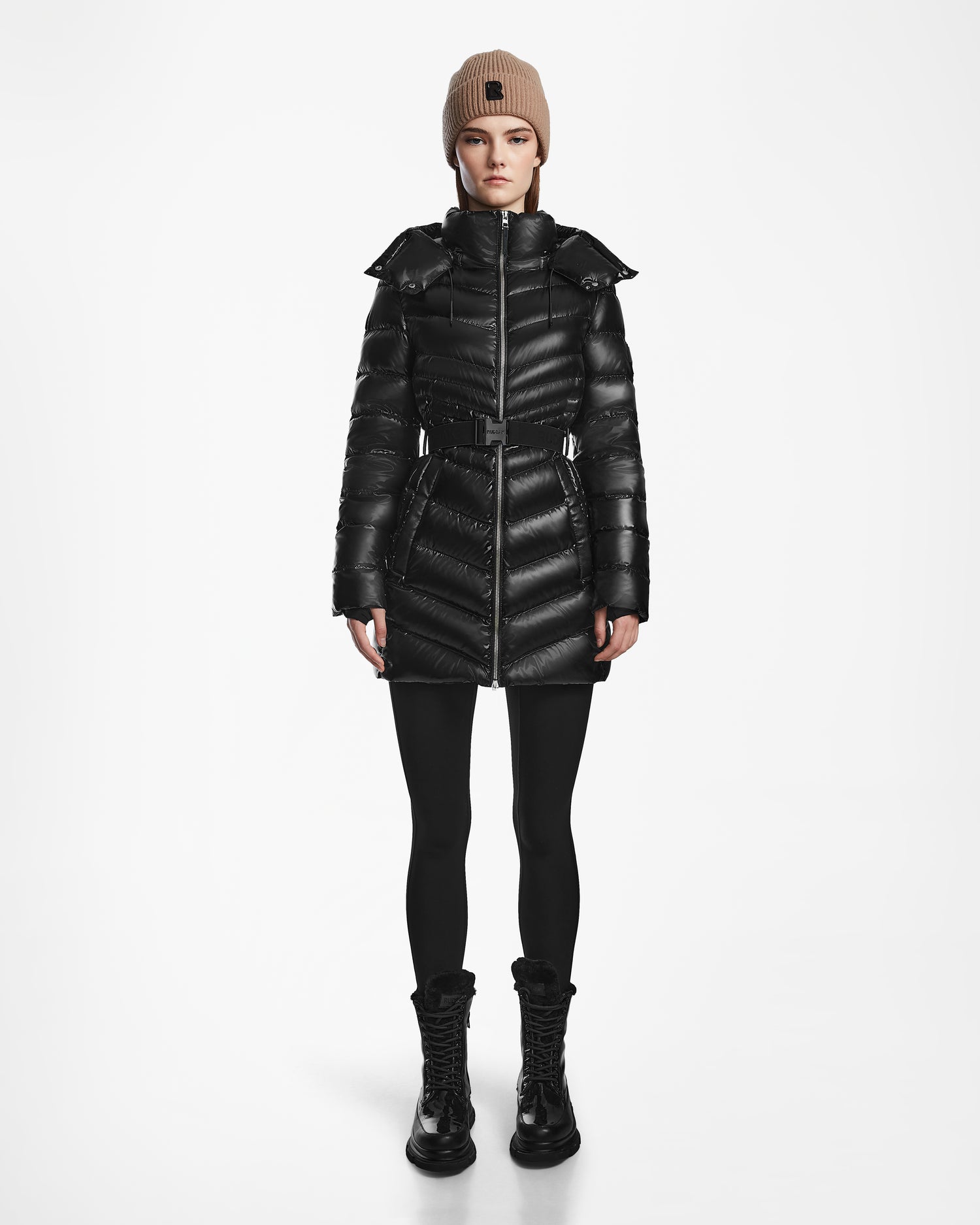 A person faces forward against a white background wearing the Rudsak MALAYA S - L123510 Black water-resistant women’s down puffer jacket, styled with black leggings, black lace-up boots, and a beige knit beanie.