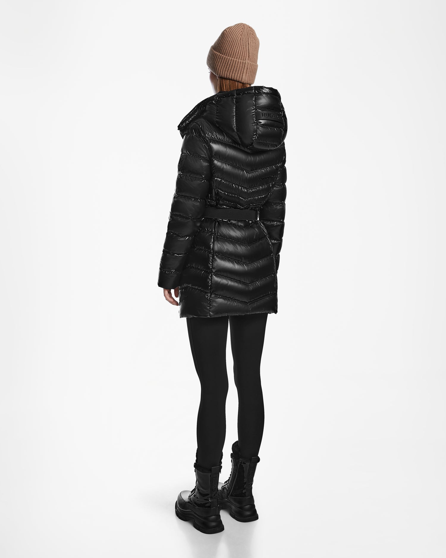 A person wears a tan beanie and the Rudsak MALAYA S - L123510 Black women’s water-resistant down puffer jacket with hood, paired with black leggings and boots, standing facing away on a plain white background.