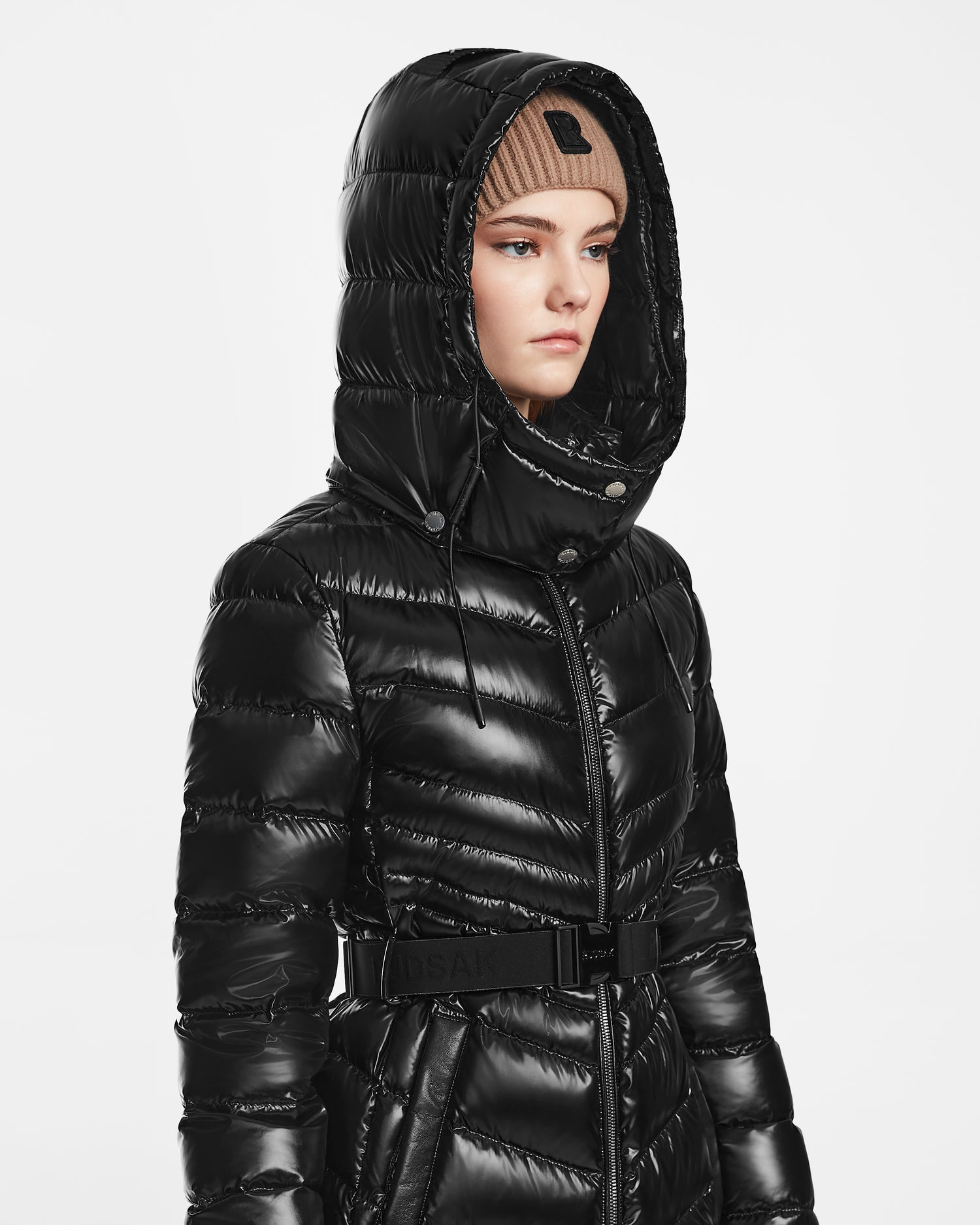A person wears the Rudsak MALAYA S - L123510 Black shiny women’s down puffer jacket with a hood and belt, paired with a light brown knit beanie, standing against a plain white background.