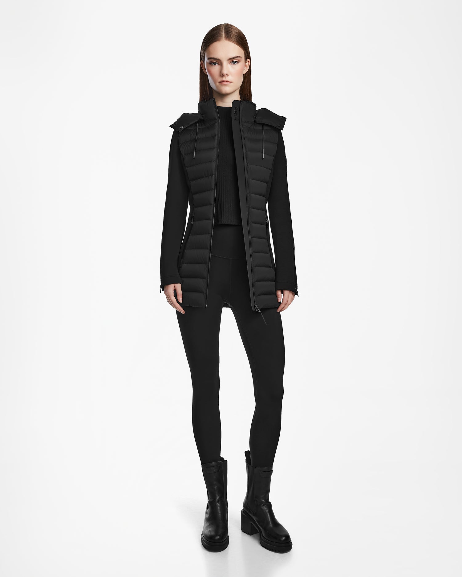 A woman stands facing forward in the MARRY M - L123548 Black puffer jacket by Rudsak, featuring a removable hood, worn over a long-sleeve top with black leggings and chunky boots against a plain white background.