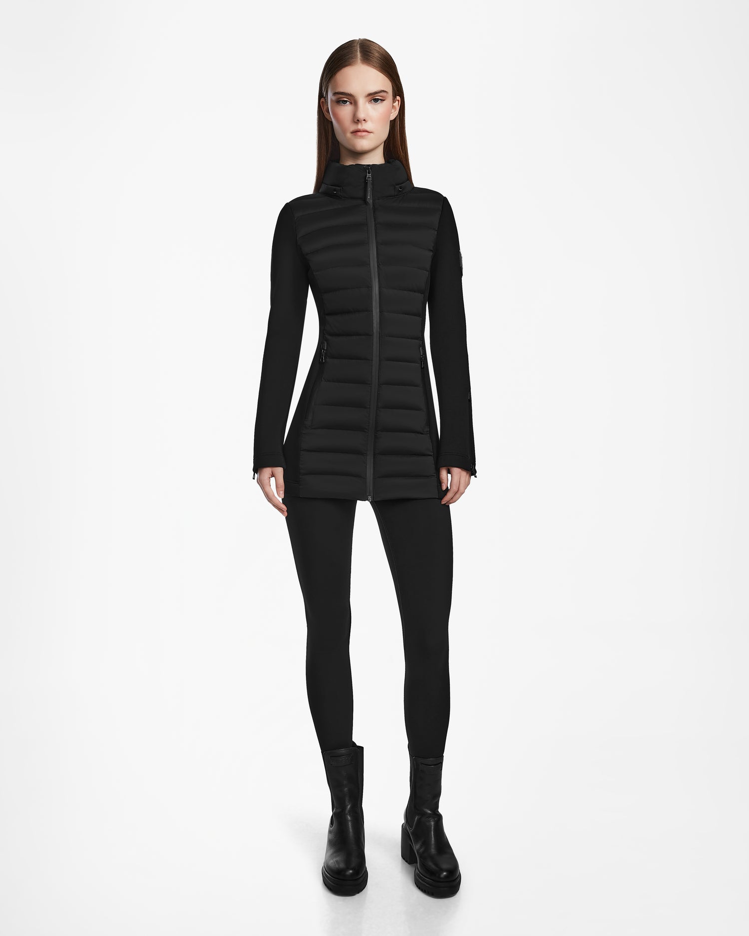 A woman stands facing forward, wearing the Rudsak MARRY M - L123548 Black women’s puffer jacket with a removable hood, paired with black leggings and boots against a plain white background.