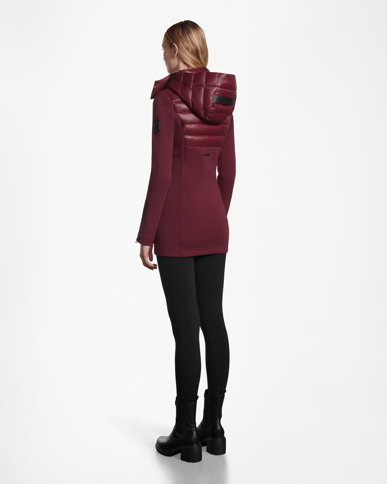 A person with blond hair faces away, wearing the Rudsak MARRY - L121003 Wine women’s puffer with padded hood, paired with black leggings and boots against a plain white background.