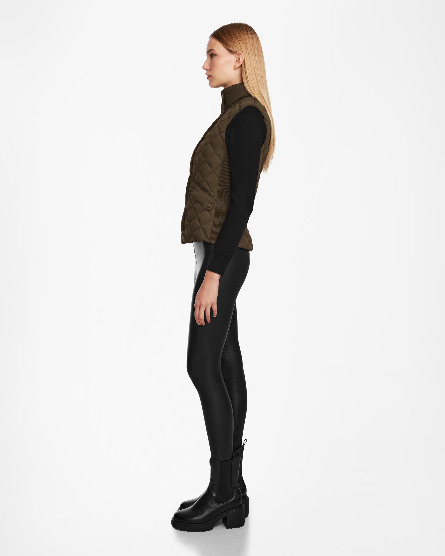 A woman with long blonde hair stands in profile against a white background, wearing the Rudsak MATILDA - L124504 Army vest over a black long-sleeve top, shiny black leggings, and black boots.