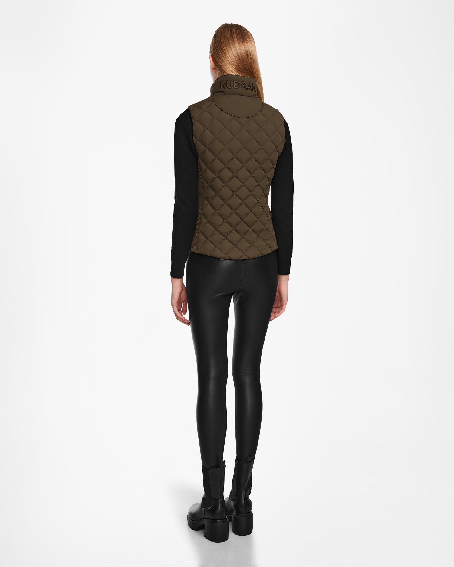 A woman with straight blonde hair faces away, wearing the Rudsak MATILDA - L124504 Army vest—a quilted dark green light layer—over a black long-sleeve shirt, leggings, and boots against a plain white background. Ideal for transitional weather.