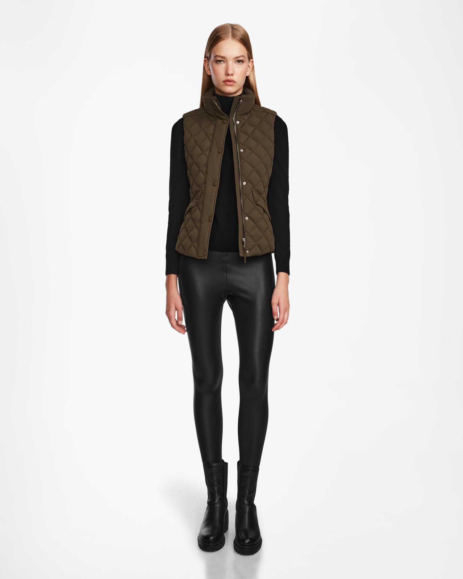 A woman faces forward against a white background, wearing the Rudsak MATILDA - L124504 Army dark green quilted vest over a black long-sleeve top, black leather-look leggings, and black ankle boots.