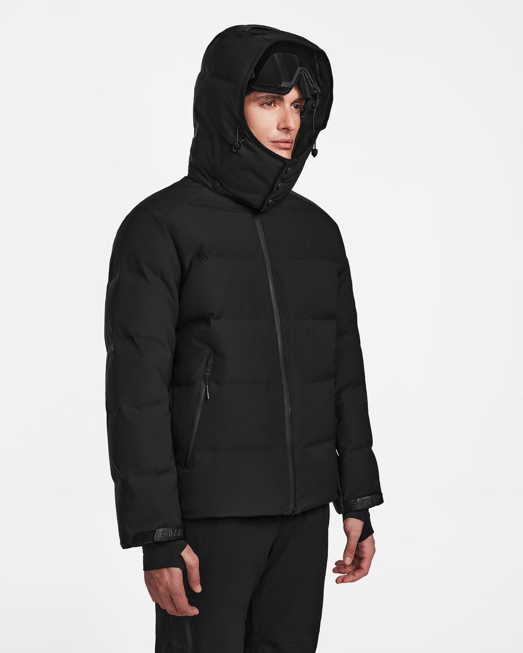 A person wearing the Rudsak MICHEL - 6124517 Black men’s ski jacket with RECCO rescue technology and black ski goggles stands facing slightly to the side against a plain white background.