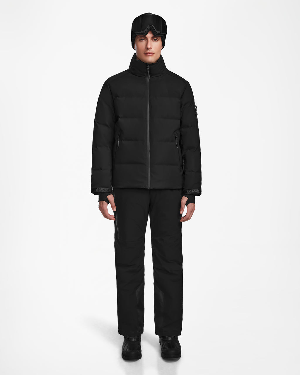 A person faces forward against a white background wearing the Rudsak MICHEL - 6124517 Black men’s ski jacket with RECCO rescue technology, black pants, boots, gloves, a beanie, and black ski goggles on their forehead.