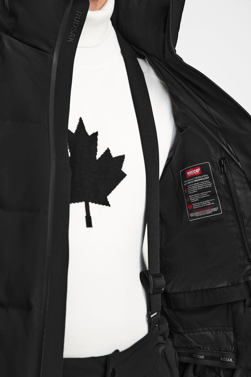 A person wears the Rudsak MICHEL - 6124517 Black men’s ski jacket, partially unzipped to reveal the label, over a white turtleneck sweater with a black maple leaf and black suspenders.