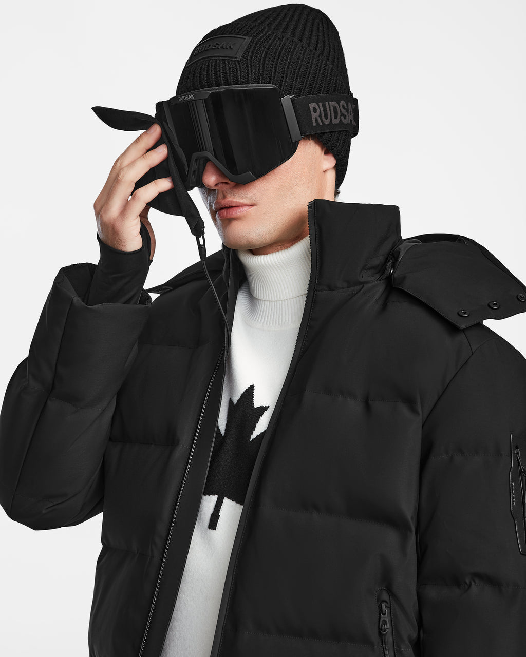 Wearing the Rudsak MICHEL - 6124517 Black men’s puffer ski jacket with RECCO technology, a black beanie, oversized black ski goggles, and a white maple leaf turtleneck, a person adjusts their goggles.