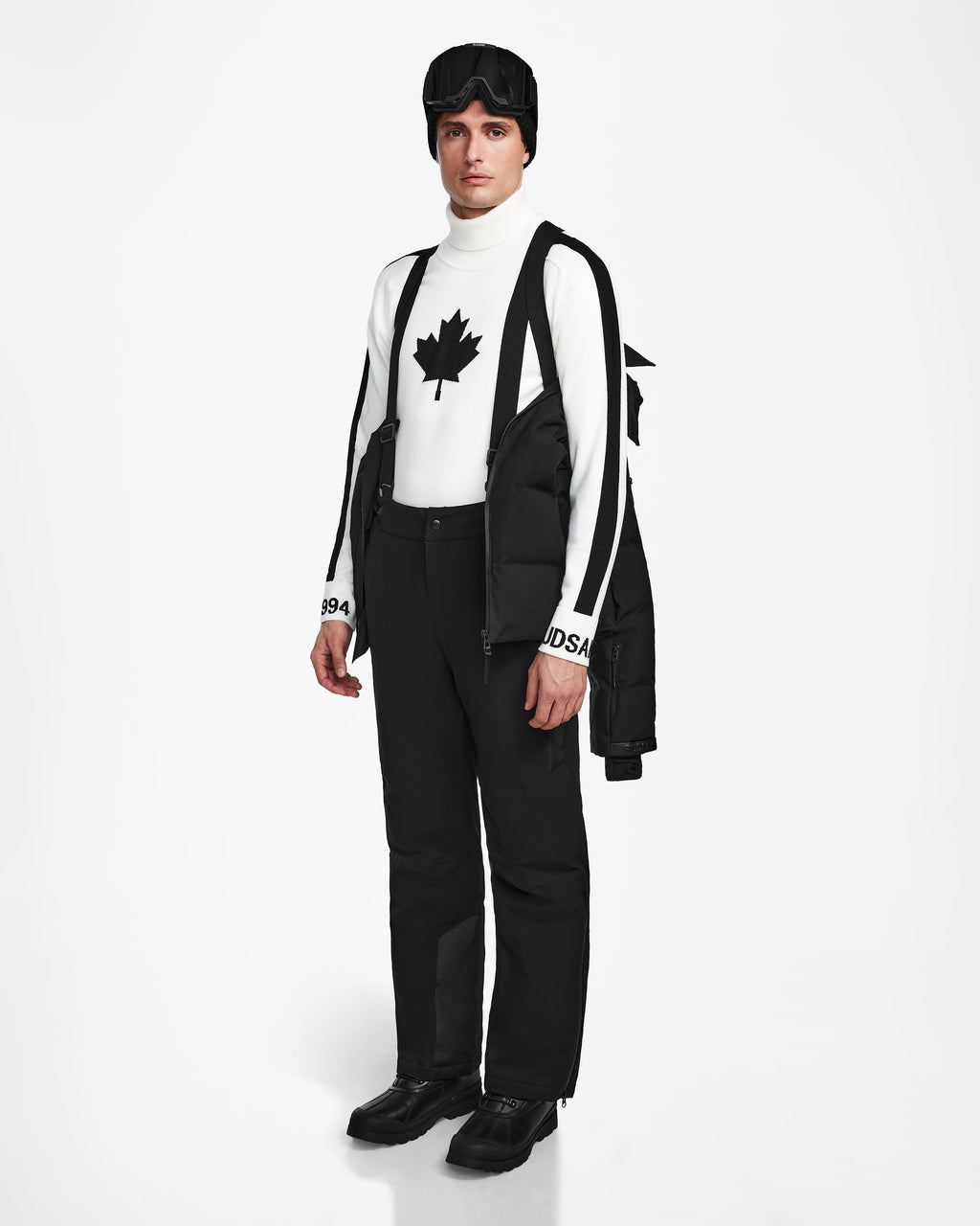 A person stands against a white background wearing Rudsaks MICHEL - 6124517 Black mens ski jacket with RECCO rescue technology over their shoulders, black snow pants and boots, a maple leaf turtleneck, suspenders, and ski goggles.