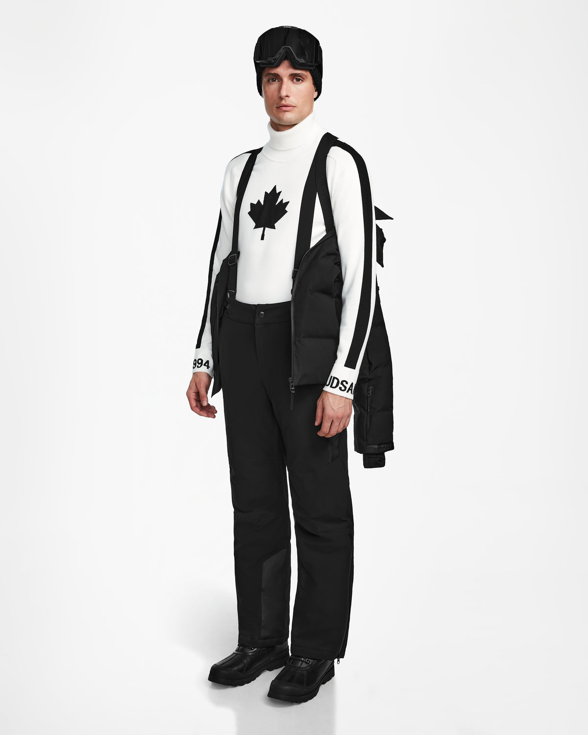A person stands against a white background wearing Rudsaks MICHEL - 6124517 Black mens ski jacket with RECCO rescue technology over their shoulders, black snow pants and boots, a maple leaf turtleneck, suspenders, and ski goggles.
