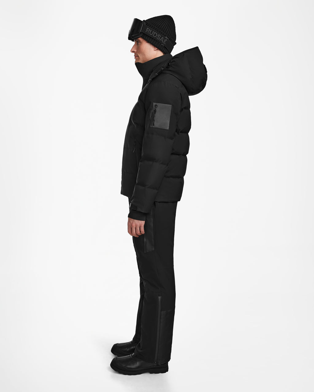A person in profile wears the Rudsak MICHEL - 6124517 Black mens ski jacket with hood, black pants, boots, beanie, and ski goggles. The jacket features RECCO rescue technology. Background is plain white.