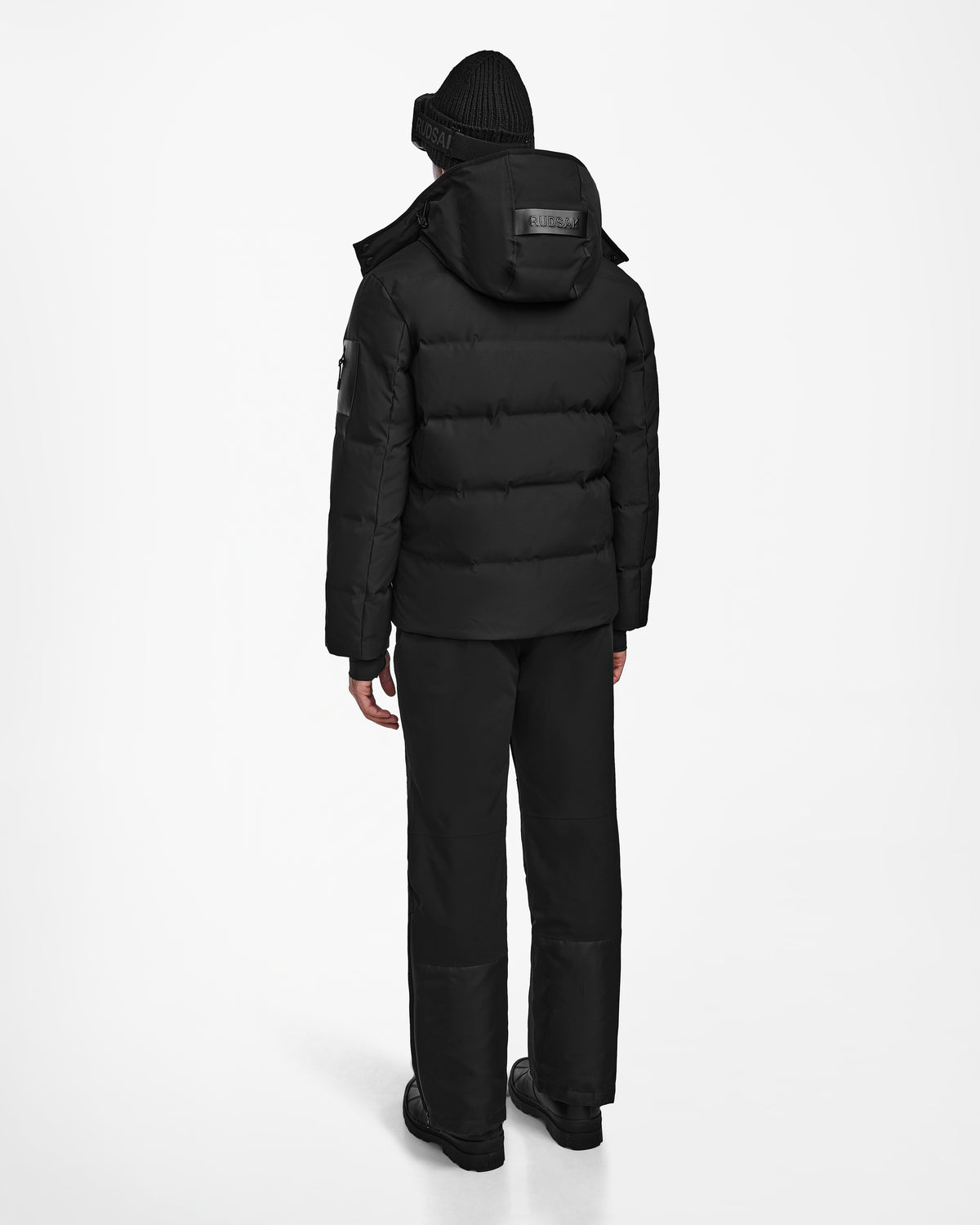 A person wearing the Rudsak MICHEL - 6124517 Black men’s ski jacket with RECCO rescue tech, along with black beanie, pants, and boots, stands facing away against a plain white background.