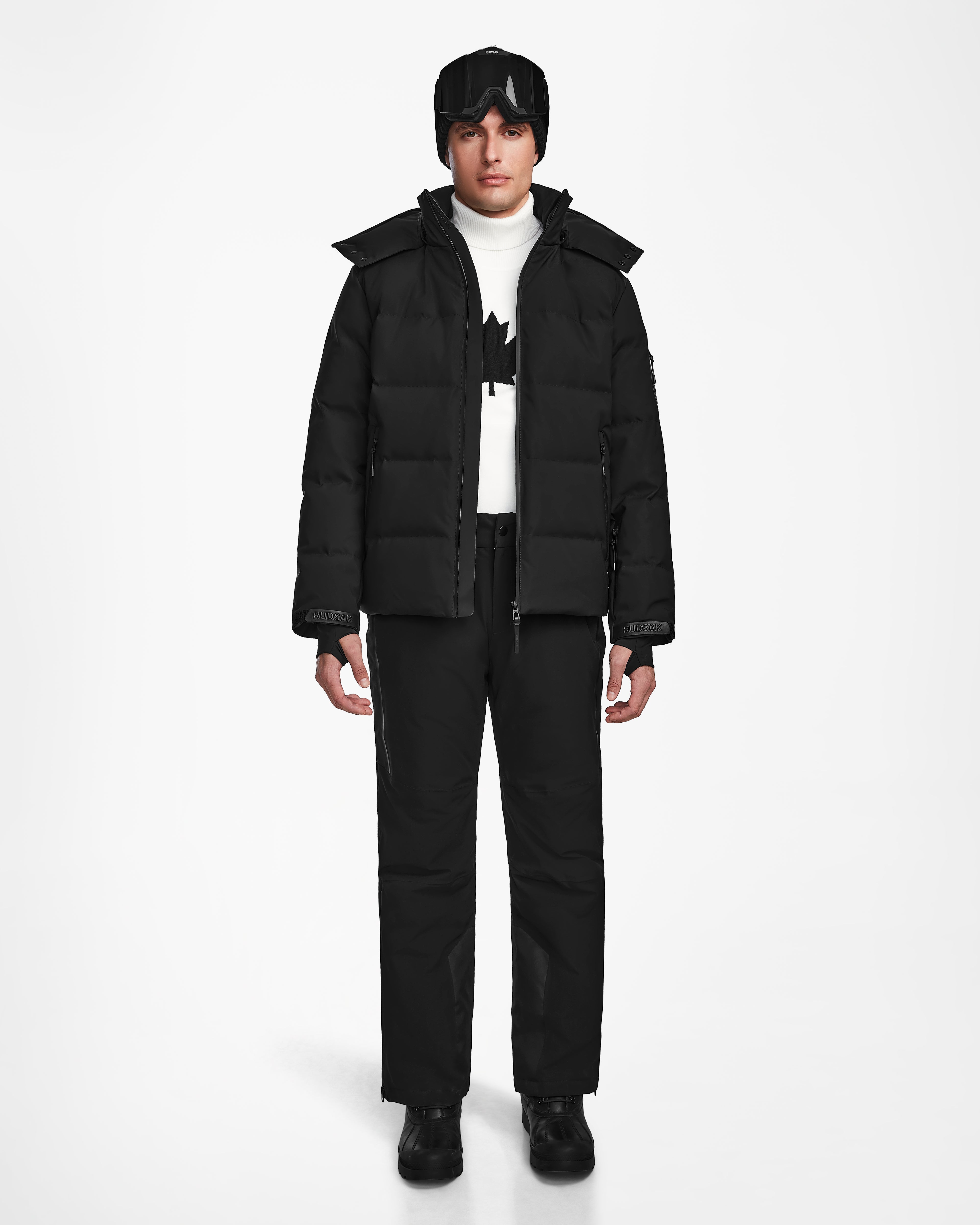 A person faces forward wearing the Rudsak MICHEL - 6124517 Black men’s ski jacket, black pants, boots, gloves, beanie, ski goggles, and a white turtleneck sweater with a black maple leaf design.