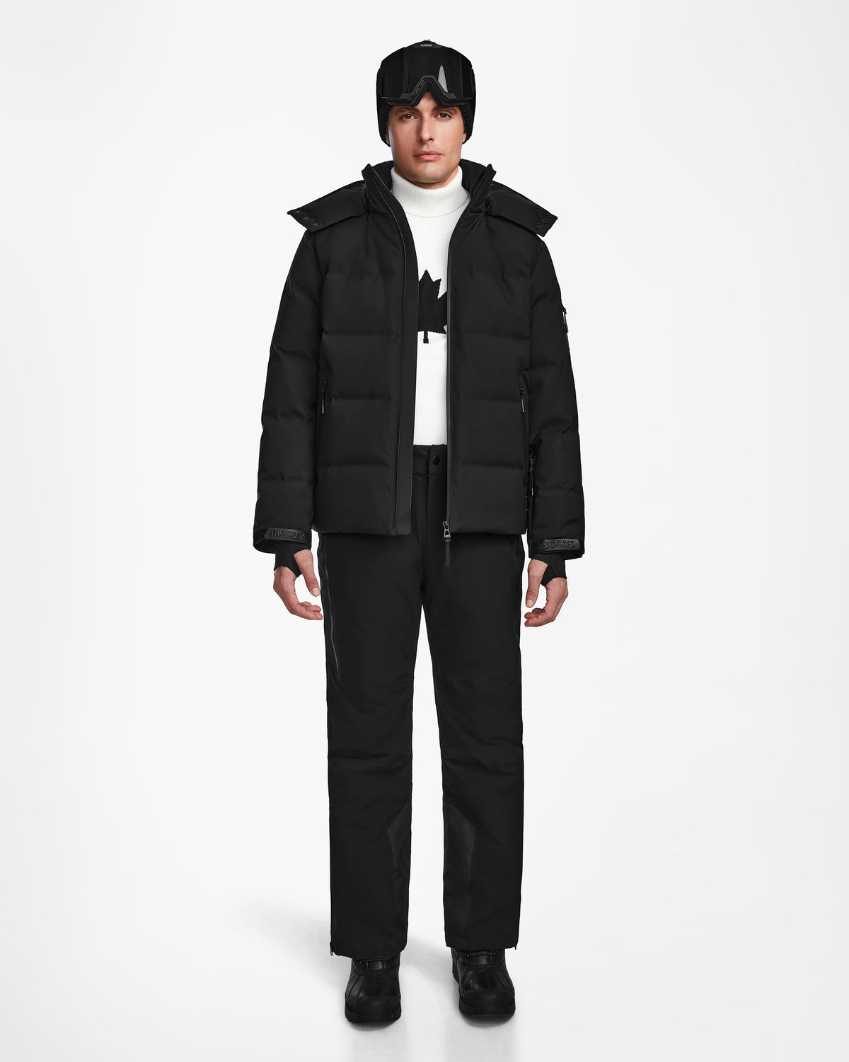 A person faces forward wearing the Rudsak MICHEL - 6124517 Black men’s ski jacket, black pants, boots, gloves, beanie, ski goggles, and a white turtleneck sweater with a black maple leaf design.