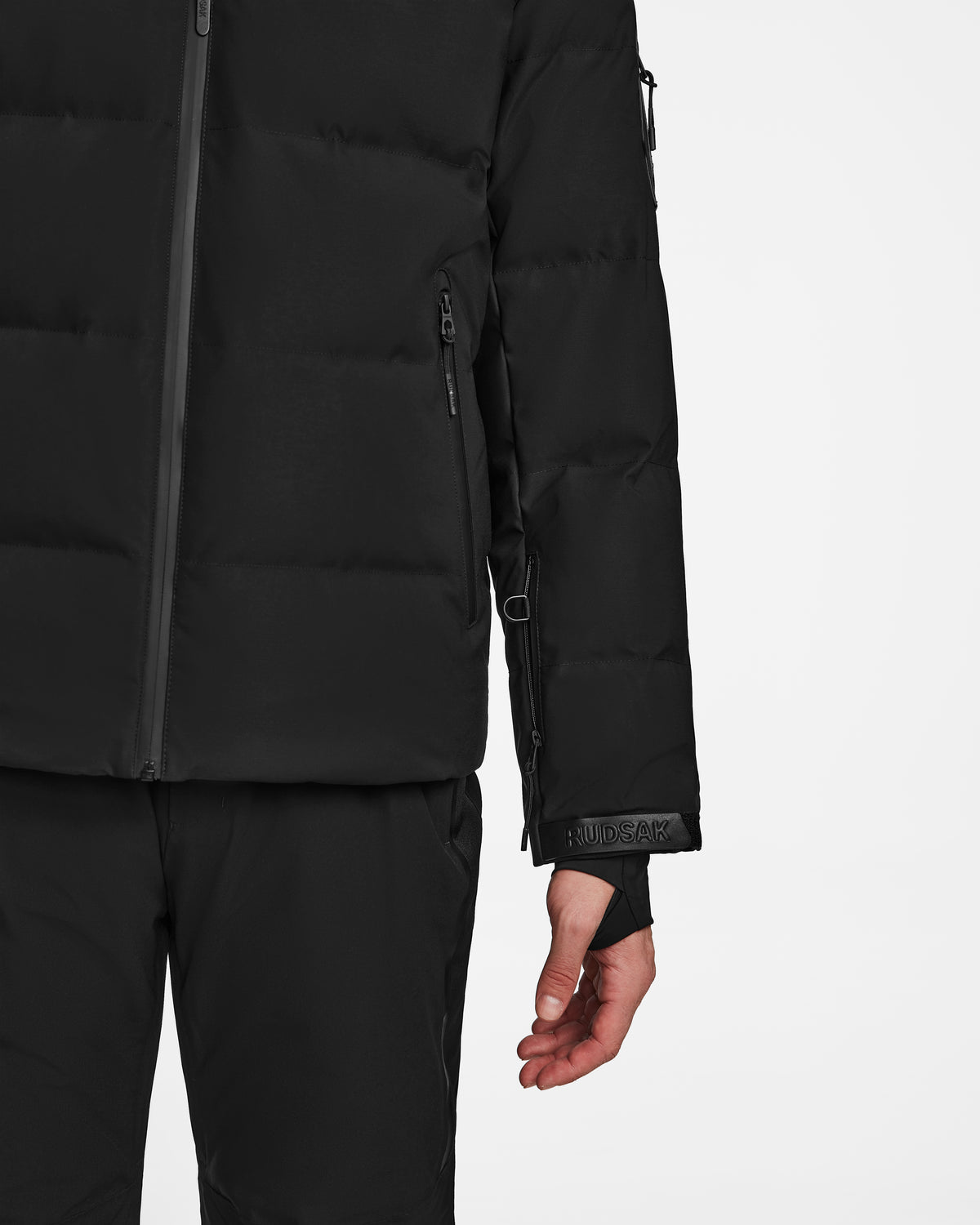 A person wearing Rudsaks MICHEL - 6124517 Black mens ski jacket with RECCO rescue technology and black pants is shown from the torso down, left arm relaxed against a plain white background.