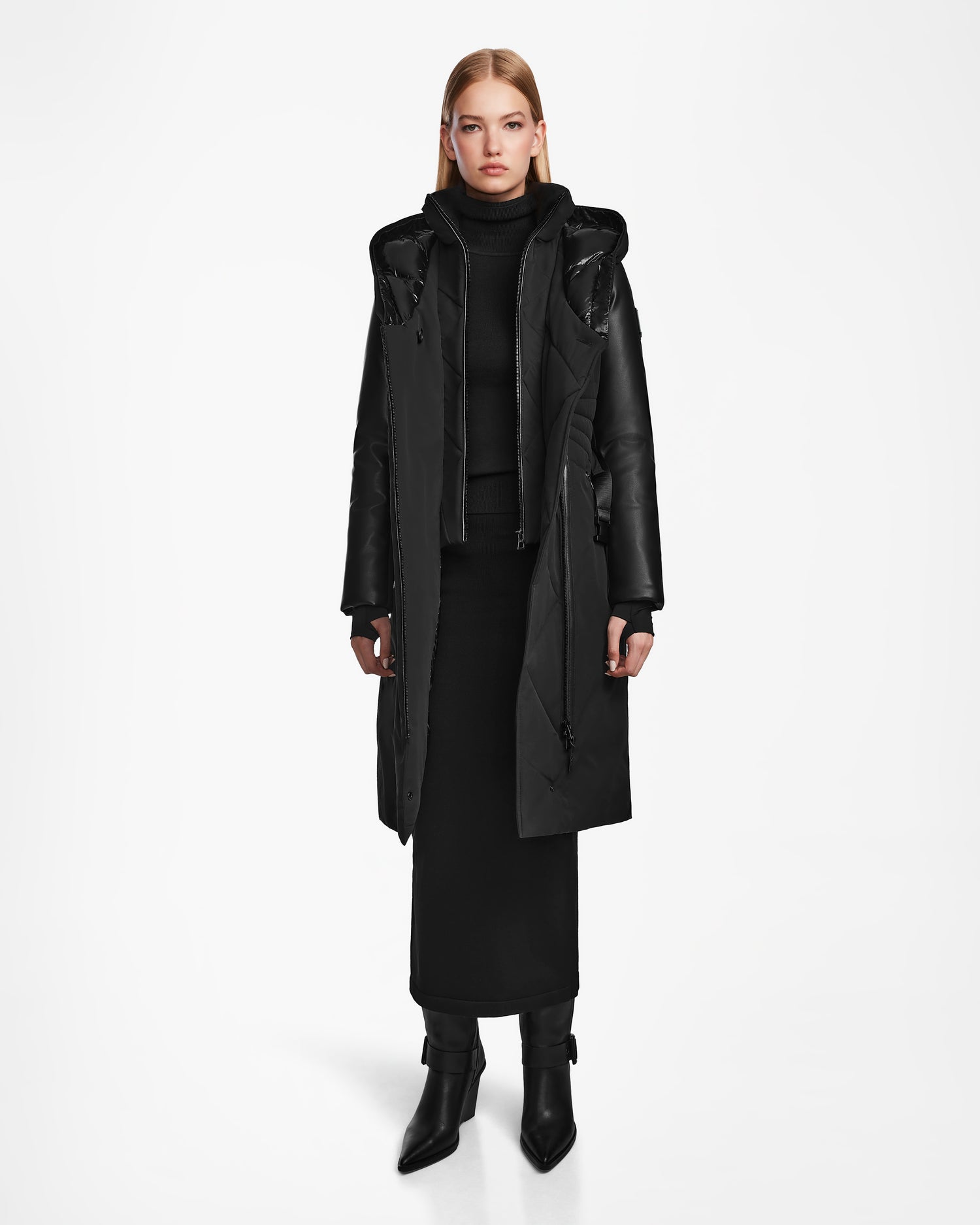A person faces forward in a plain white setting, wearing the Rudsak MISALA - L124512 Black women’s long down coat over a black turtleneck top, long skirt, and black boots.