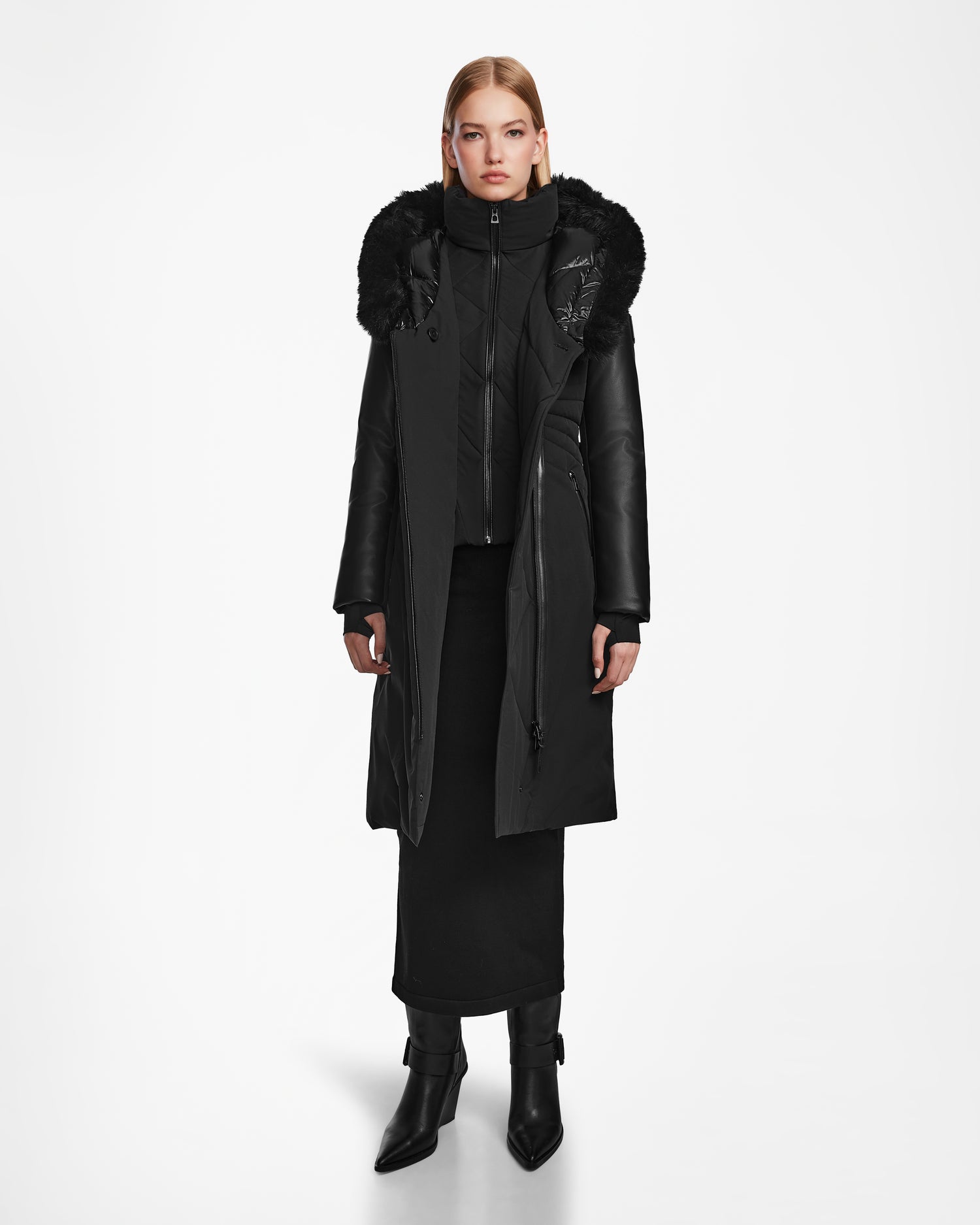 A woman faces forward wearing the Rudsak MISALA - L124512 Black long down coat with a fur-trimmed hood, paired with a black skirt, high-neck inner layer, and black heeled boots against a plain white background.