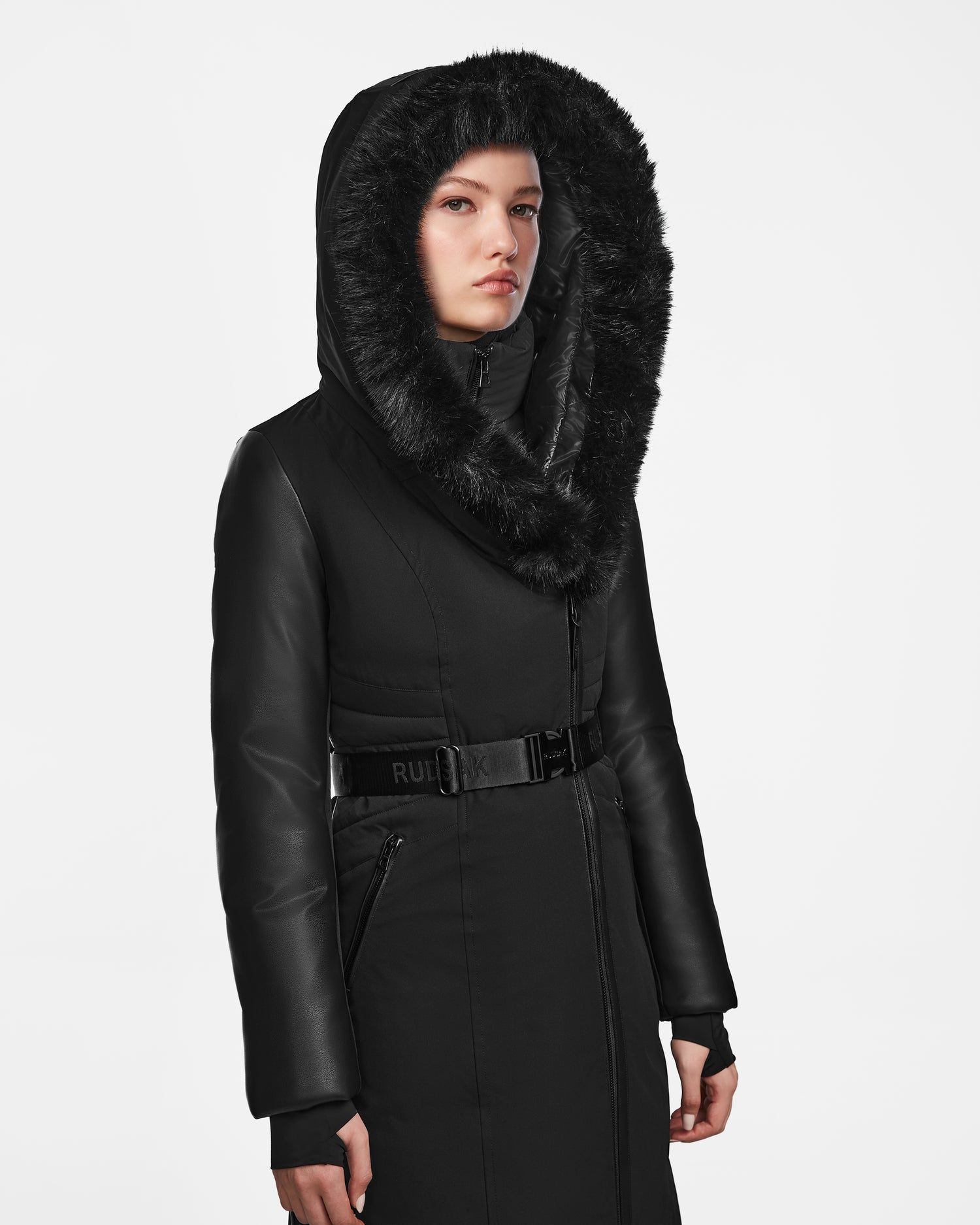 A woman models the Rudsak MISALA - L124512 Black womens long down coat with a fur-lined hood, belted waist, and leather-like sleeves. The water-resistant winter coat is showcased against a plain white background.