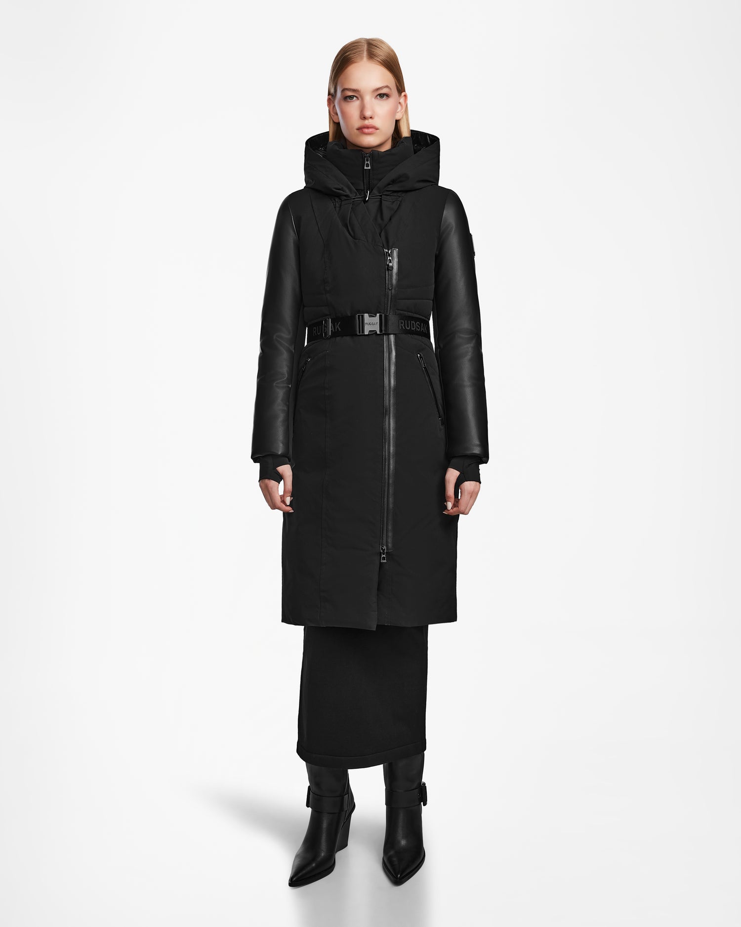 A woman faces forward in the Rudsak MISALA - L124512 Black water-resistant winter coat with a high collar and hood, paired with a black skirt and heeled ankle boots, against a plain white background.