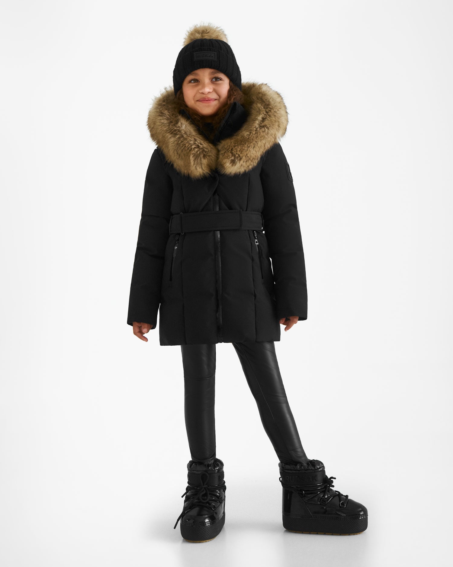 A young girl in a black Rudsak MODA K - 0124505 winter coat with a fur-lined hood, matching beanie, faux leather pants, and boots smiles against a white background. The parka is made from recycled materials.