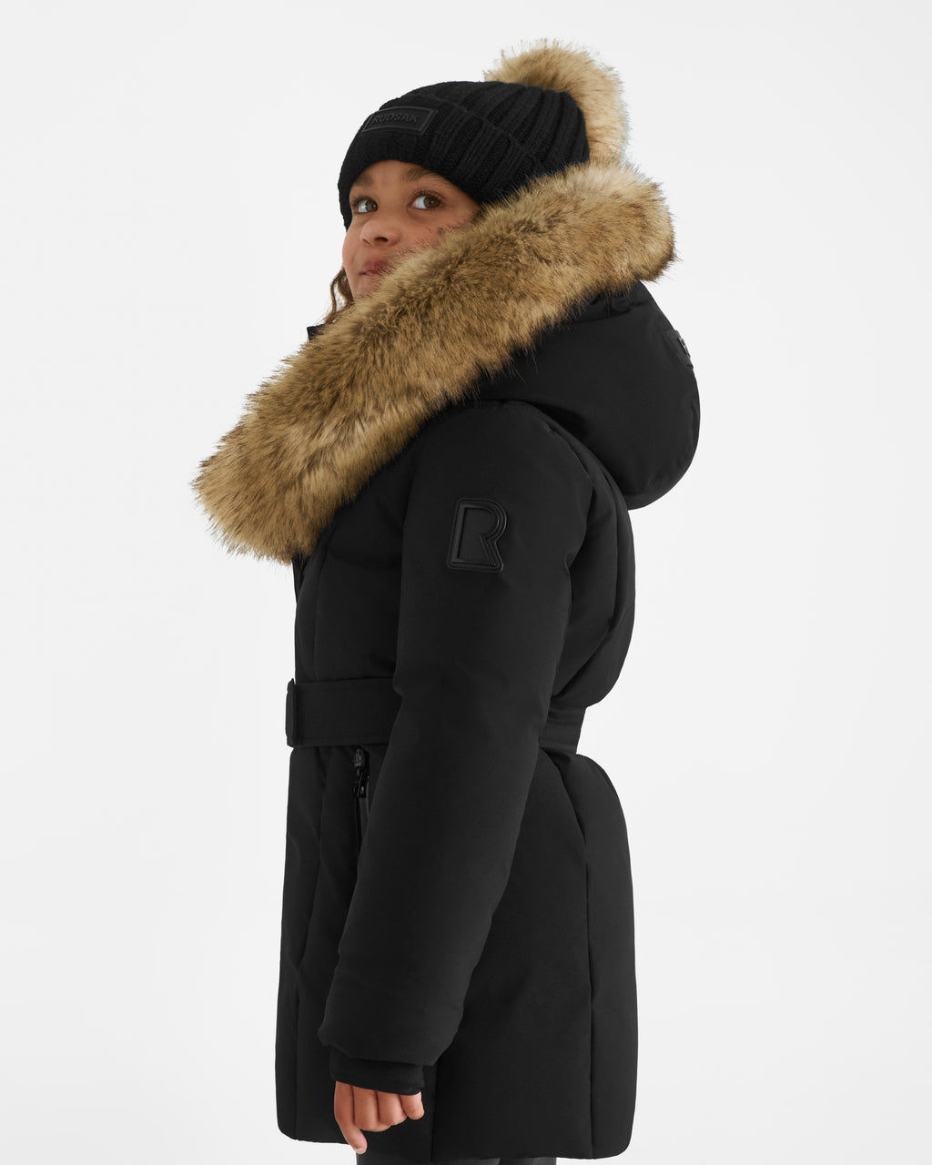 A young child in Rudsak’s MODA K - 0124505 Black down parka with a fur-lined hood and matching knit hat stands sideways, looking up against a white background; the winter coat is made from recycled materials.
