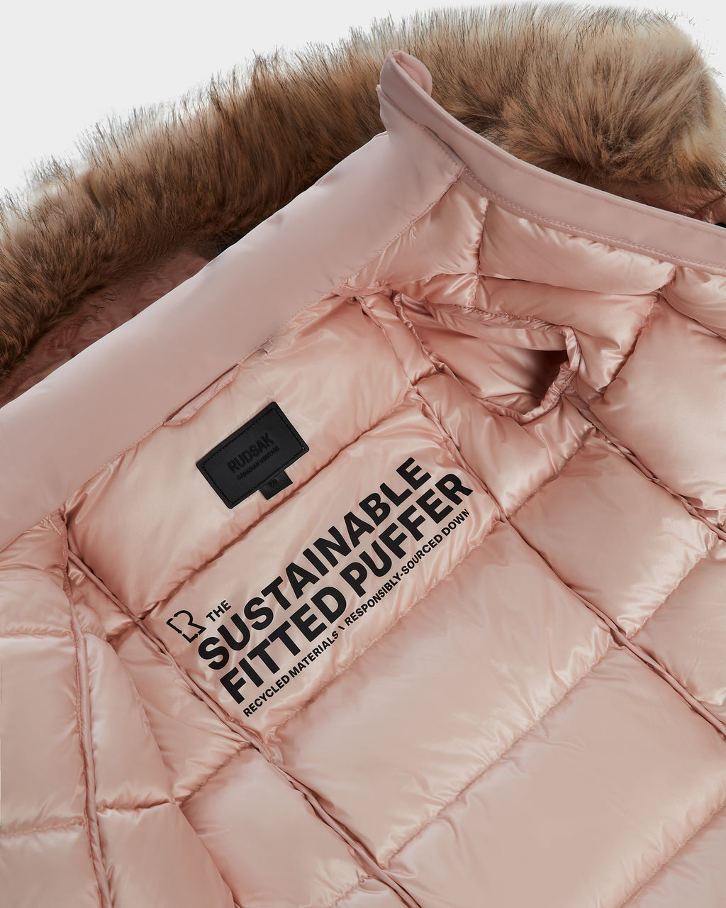 A close-up of the Rudsak MODA K - 0124505 Pastel Pink girl's down parka with a faux fur-trimmed hood. The label, THE SUSTAINABLE FITTED PUFFER, highlights recycled nylon and responsibly sourced down.