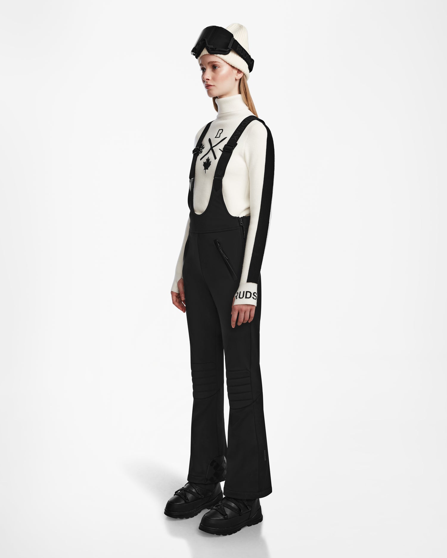 A person wears Rudsaks NANOR - 8124819 Black fitted flared womens ski pants, paired with a white turtleneck featuring black accents, black gloves, ski goggles, and a white hat, standing left against a plain white background.