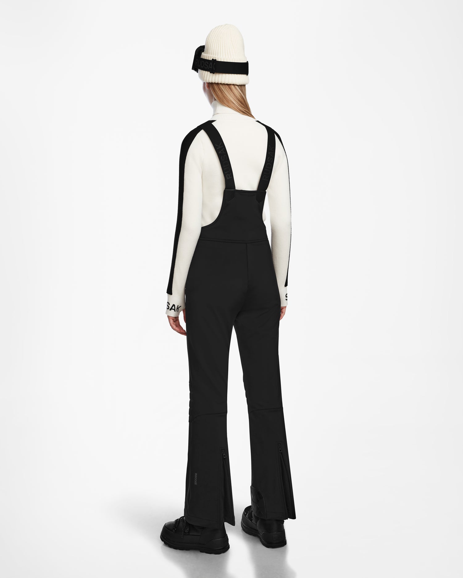 A person facing away, wearing Rudsaks NANOR - 8124819 Black fitted flared ski pants with a white turtleneck, a white beanie with a black stripe, and black shoes, stands against a plain white background.