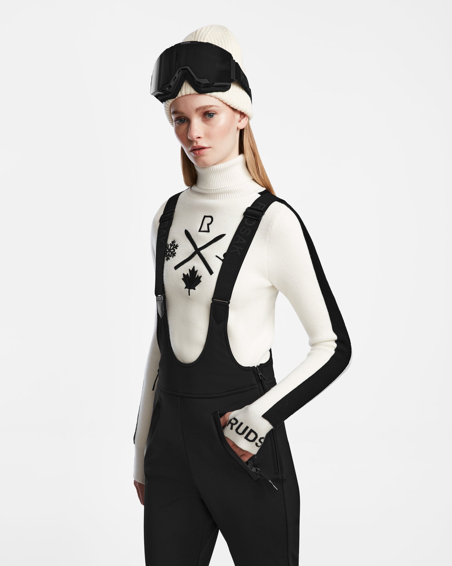 A woman in Rudsak’s NANOR - 8124819 Black flared ski pant overalls, a white turtleneck with black winter graphics, cream knit beanie, and ski goggles stands against a plain white background.