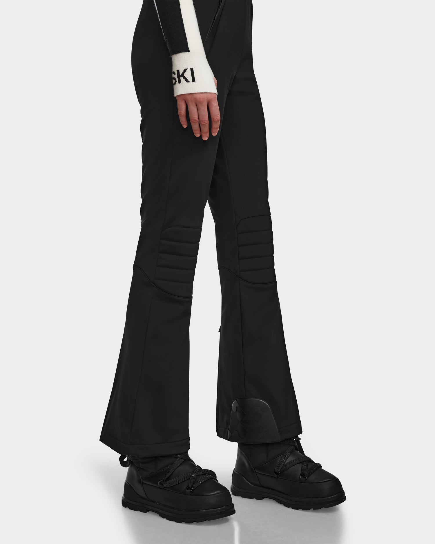 A person wearing Rudsak NANOR - 8124819 Black fitted flared ski pants and black snow boots stands against a plain white background. Only the lower half is visible, with one hand in a black-and-white SKI glove at their side.