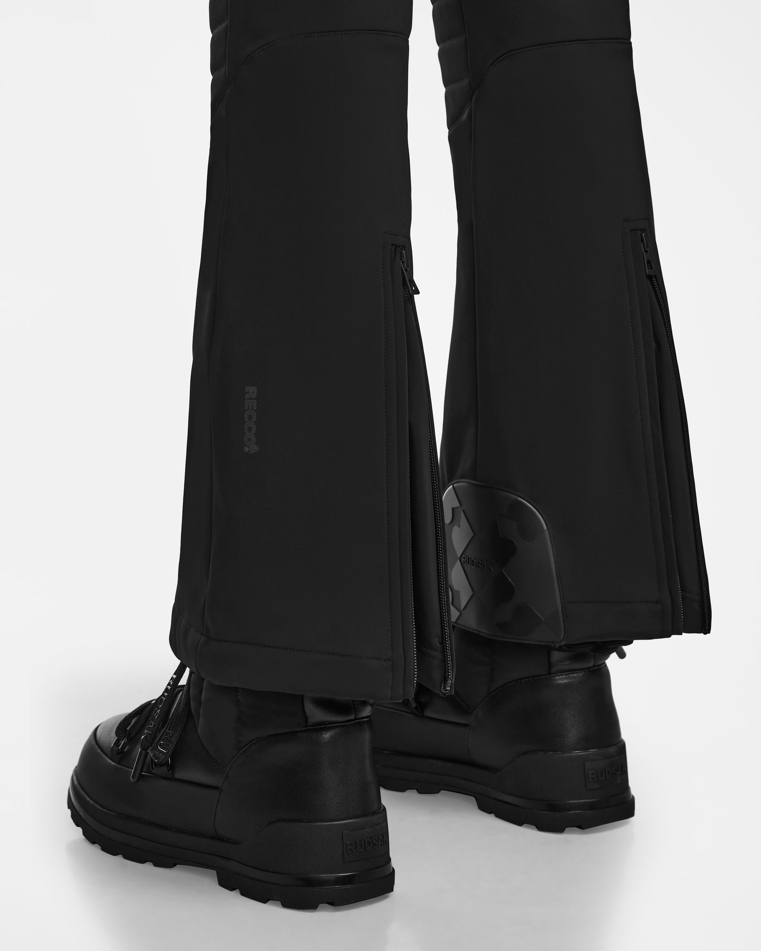 Close-up of a person wearing Rudsak NANOR - 8124819 Black womens fitted flared ski pants with visible branding, back zippers, RECCO rescue technology, and a small ankle detail, paired with black boots. Photographed on a white background.