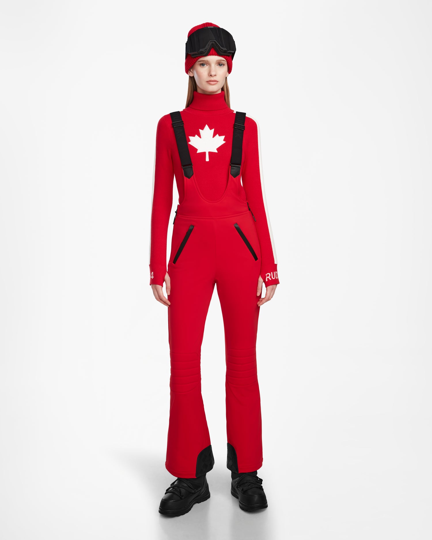 A person faces forward in the Rudsak NANOR - 8124819 Flame Red ski outfit with black accents, fitted flared ski pants, a red top featuring a white maple leaf, a matching hat, black shoes, and RECCO rescue tech against a white background.