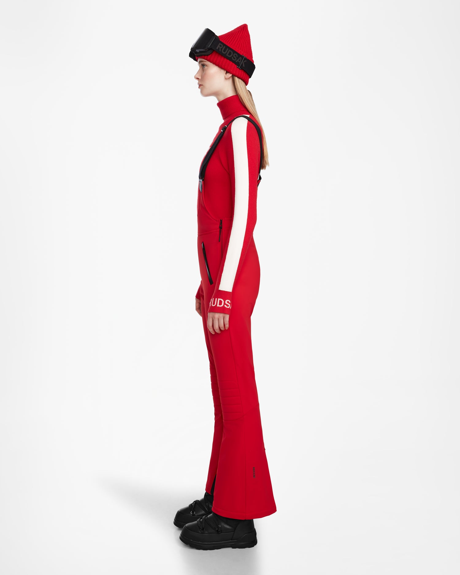 A woman stands in profile against a white background, wearing Rudsaks NANOR - 8124819 Flame Red fitted flared ski pants and a matching beanie, paired with black boots and her hands relaxed at her sides.