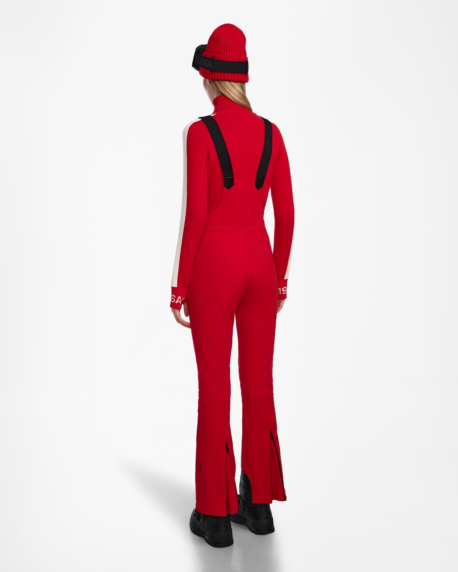 A person is seen from behind in the Rudsak NANOR - 8124819 Flame Red ski suit, featuring black suspenders, fitted flared pants with back zippers, white sleeve accents, a red and black knit hat, and black boots.