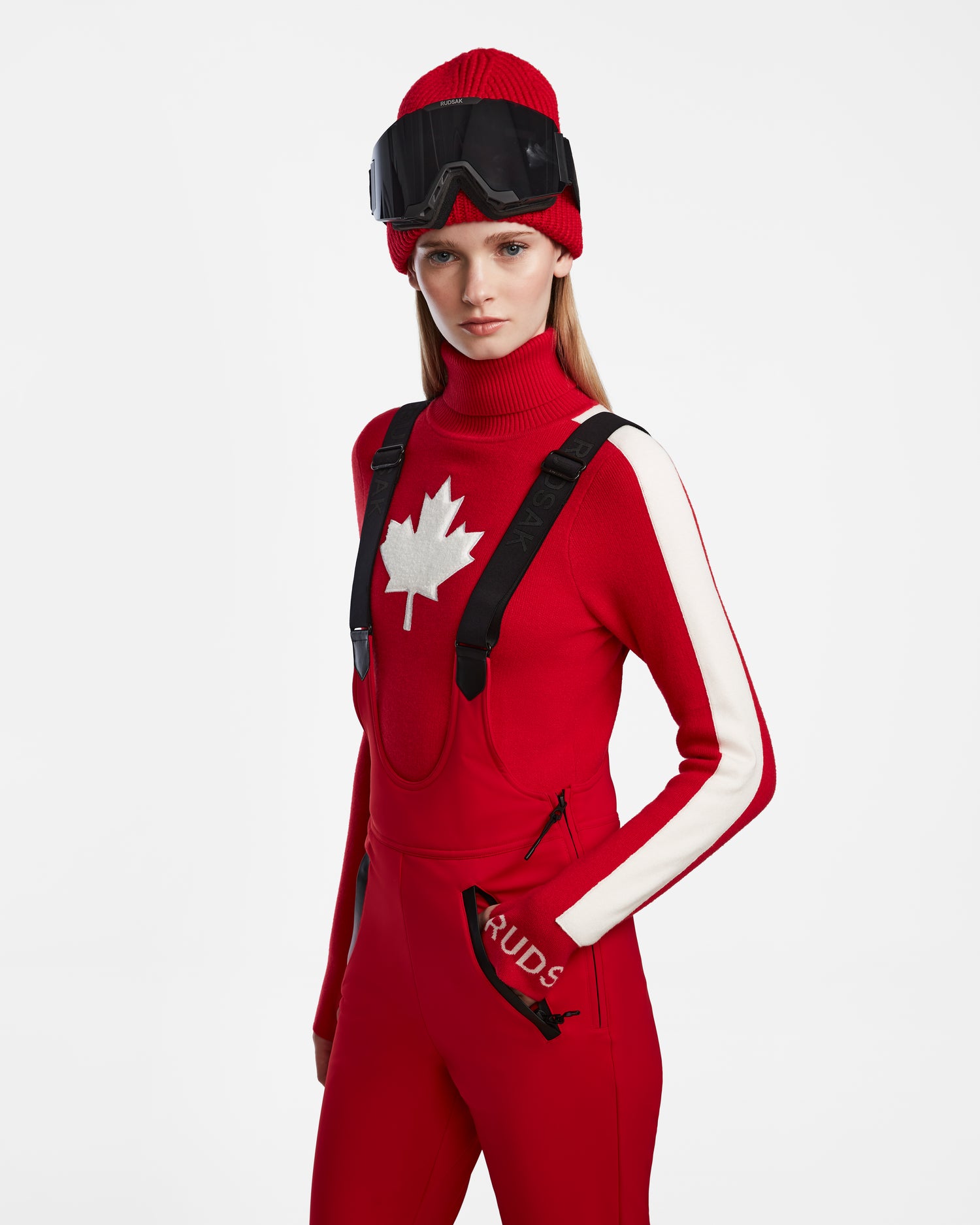 A woman in a red knit hat and ski goggles wears the Rudsak NANOR - 8124819 Flame Red fitted flared womens ski pants with RECCO rescue tech, paired with a red and white maple leaf sweater, standing against a white background.