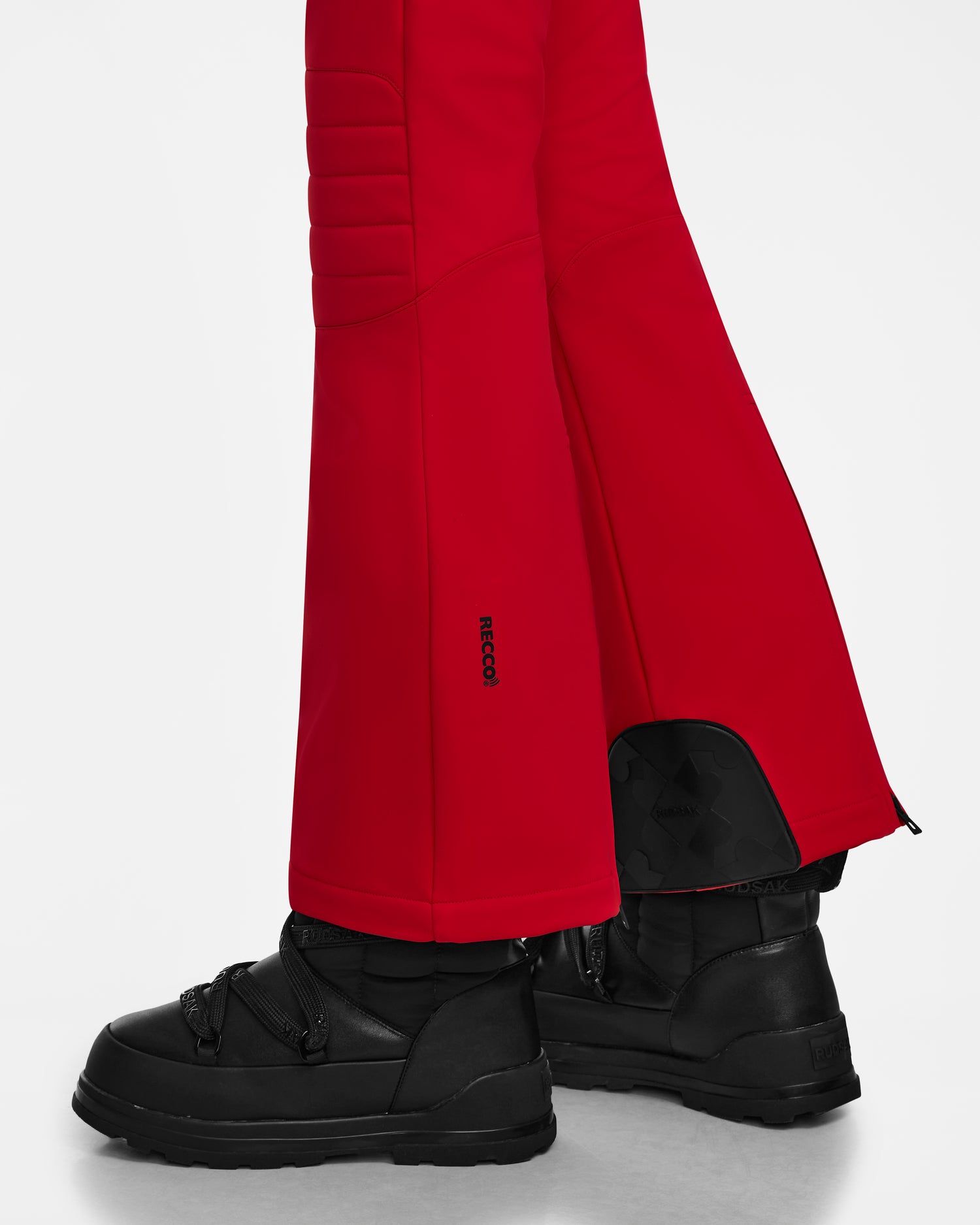 A person wearing Rudsak NANOR - 8124819 Flame Red fitted flared ski pants with padded knees and ankle zippers, paired with black velcro-strap snow boots, stands against a white background.