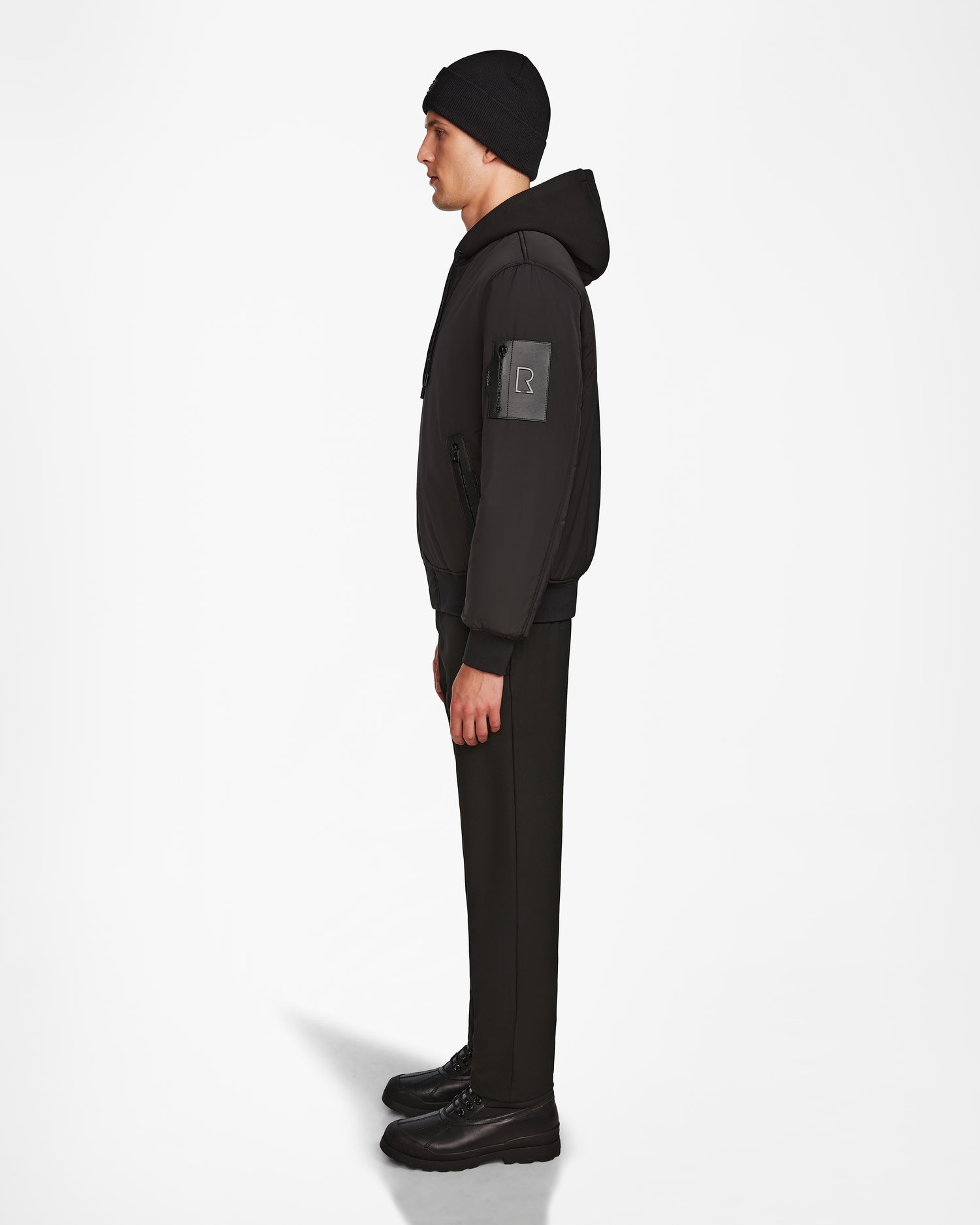 A man in profile faces left, wearing Rudsaks NEVEN - M124520 Black mens bomber jacket with a sleeve patch made from recycled materials, paired with a black beanie, pants, and shoes against a plain white background.