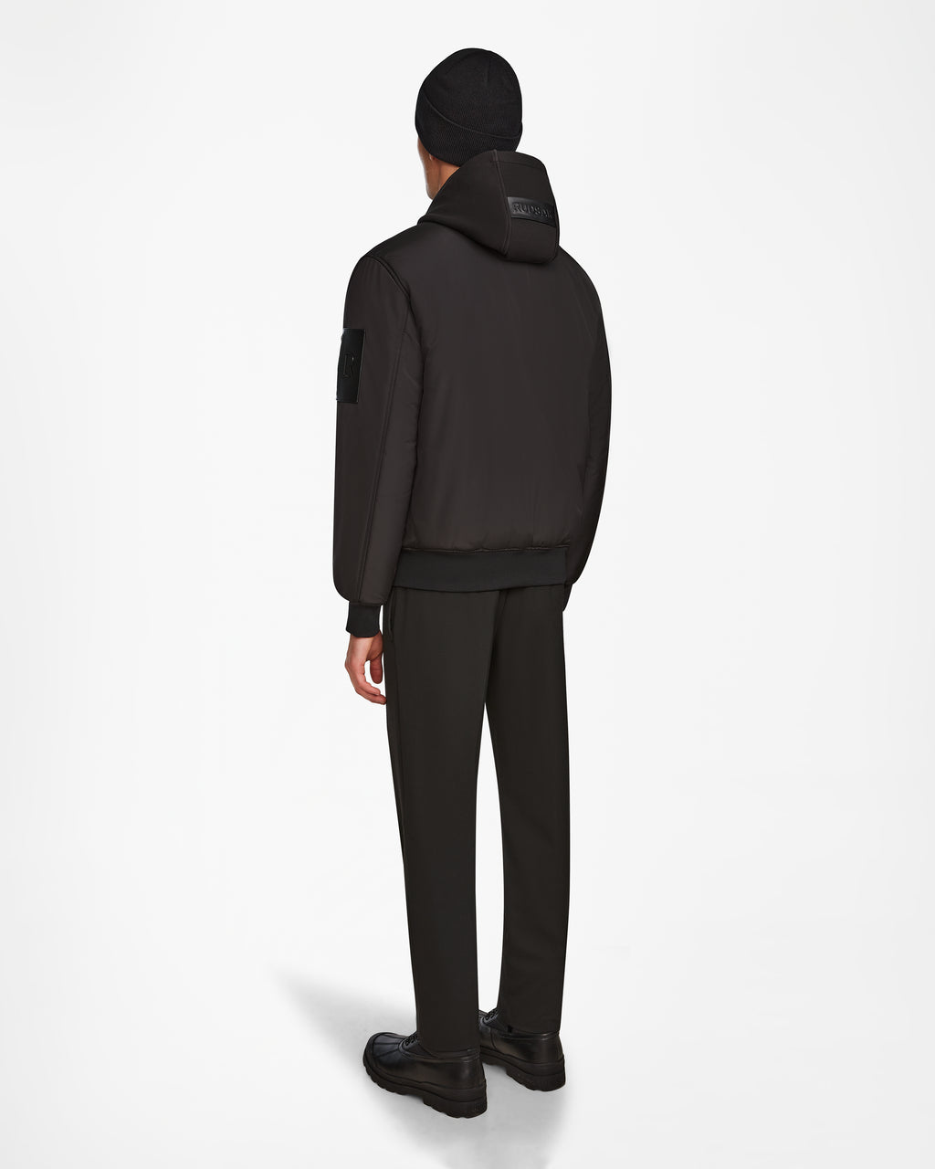 Wearing the Rudsak NEVEN - M124520 Black men’s bomber jacket, a person stands facing away in dark dress pants, black beanie, and black shoes against a white background.