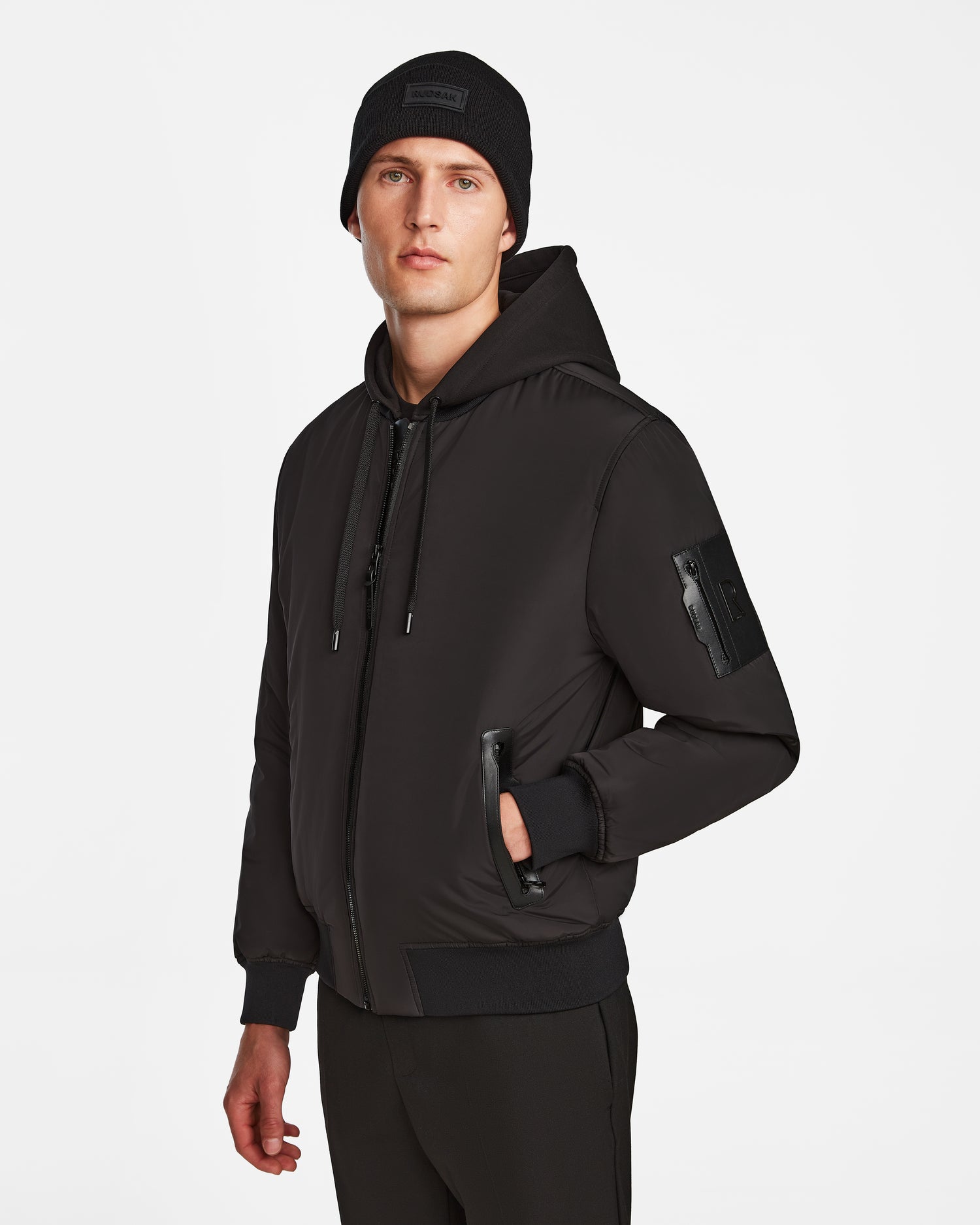 A man wearing the Rudsak NEVEN - M124520 Black men’s bomber jacket, made from recycled materials with zip closure and front pockets, black beanie, and black pants stands against a plain white background.