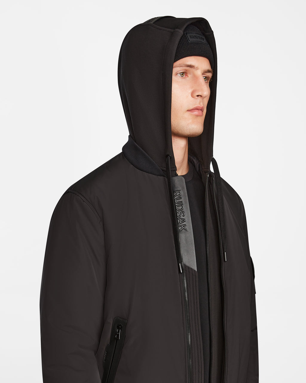 A person wears the Rudsak NEVEN - M124520 Black mens bomber jacket with a matching beanie, looking to the side against a plain white background.