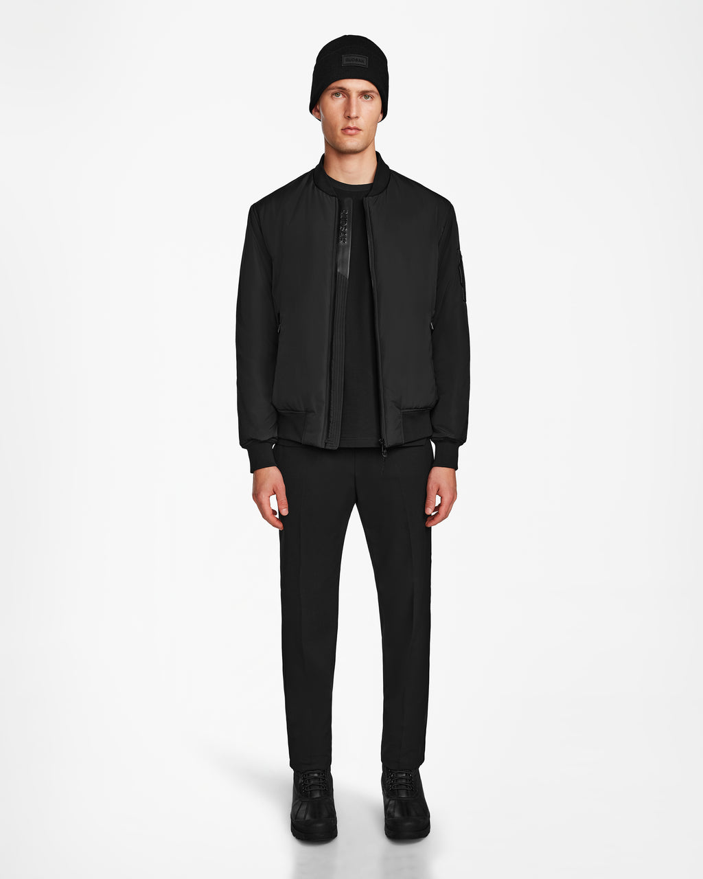 A man faces forward in a Rudsak NEVEN - M124520 Black mens bomber jacket made from recycled materials, styled with a black beanie, shirt, pants, and sneakers against a plain white background.