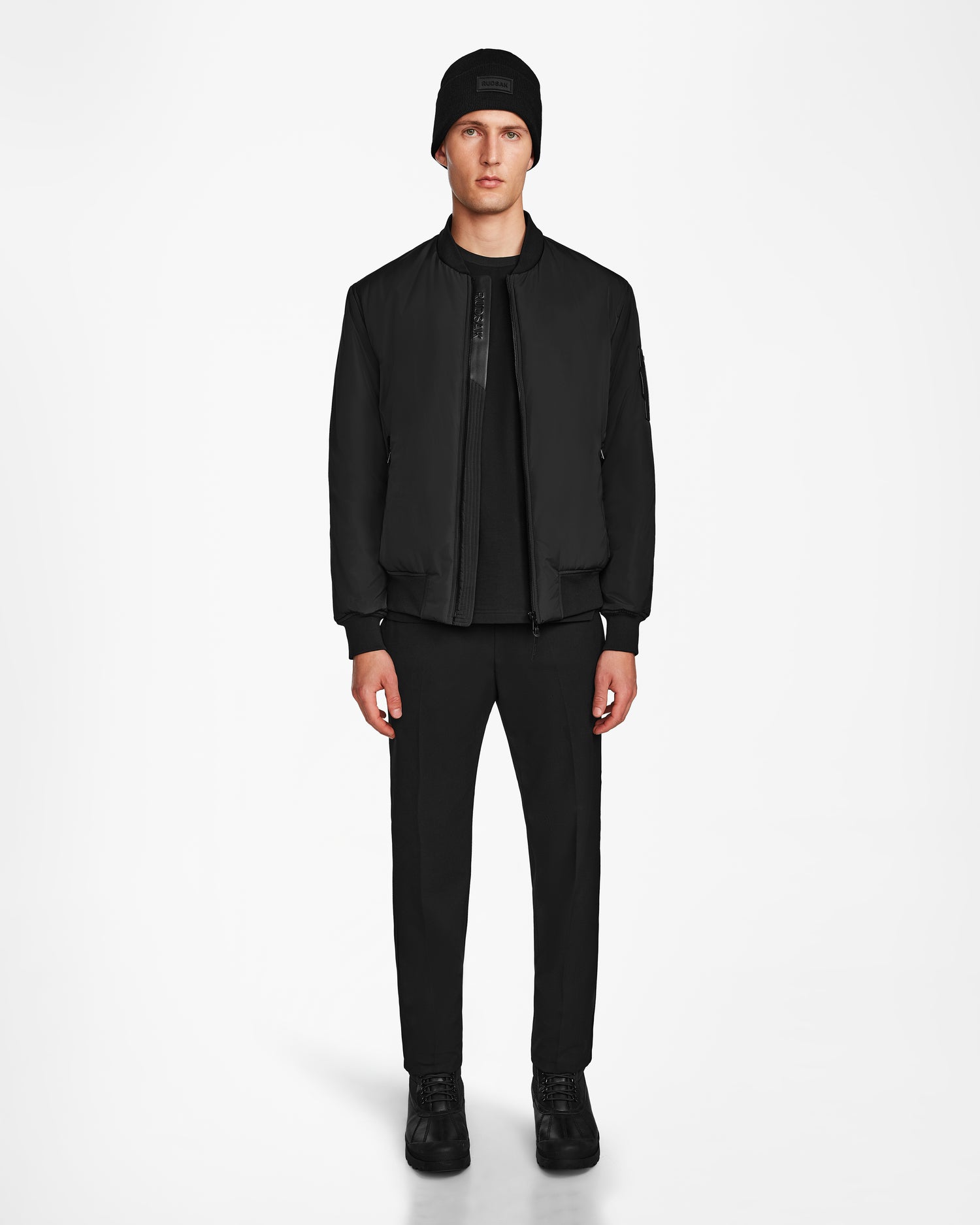 A man faces forward in a Rudsak NEVEN - M124520 Black mens bomber jacket made from recycled materials, styled with a black beanie, shirt, pants, and sneakers against a plain white background.