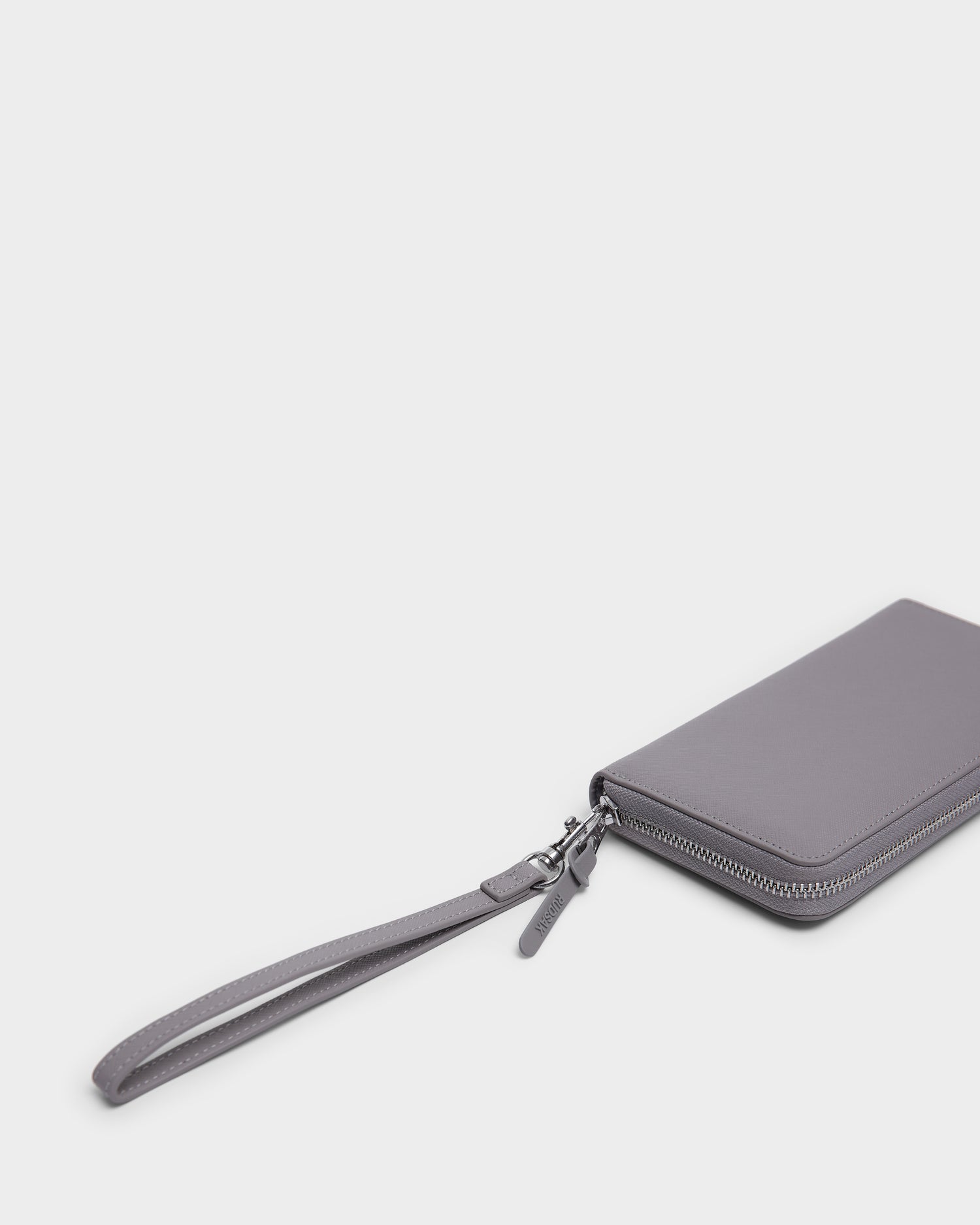 The Rudsak NYLA - L424502 Grey women’s leather wallet with a wrist strap and zippered compartment rests on a white background, with only part of the wallet and strap visible in the lower right corner.