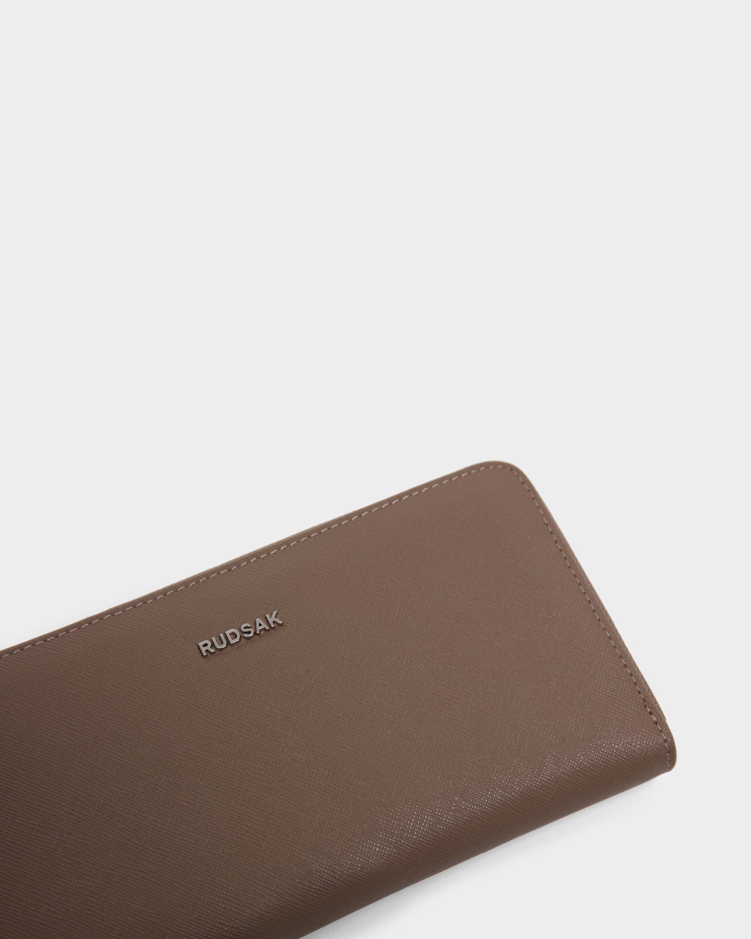 The NYLA - L424502 Sand is a brown womens leather wallet by Rudsak, with cardholder slots, a zippered compartment, and the brand name in silver lettering, shown against a plain white background.