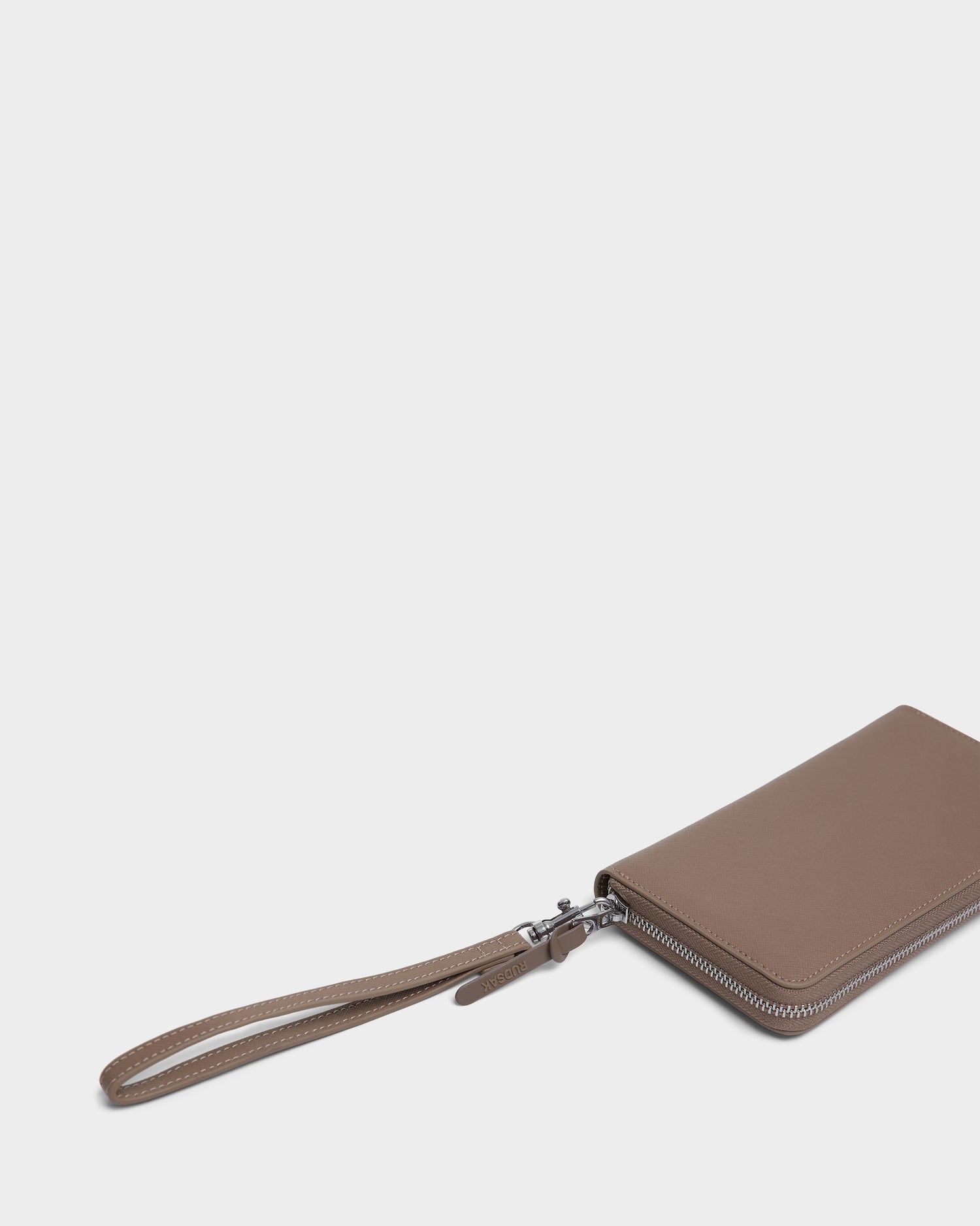 The Rudsak NYLA - L424502 Sand womens leather wallet, featuring a zippered compartment and card slots, is partially visible at the lower right corner with its brown surface and wrist strap against a plain white background.