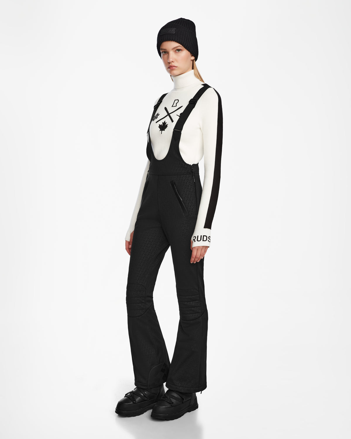A woman models the Rudsak OKSANA - 8124527 Monogram Print Black ski pants, paired with a black beanie and white turtleneck, standing against a plain white background with her hands by her sides, gazing slightly left.