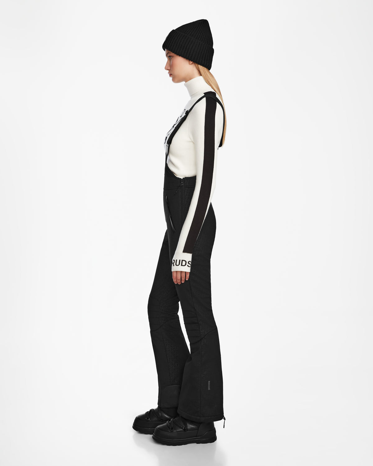A person stands in profile against a white background, wearing the Rudsak OKSANA - 8124527 Monogram Print Black flared ski pant overalls with monogram on the cuff, a black beanie, white long-sleeve top, chunky black boots, and hair in a low ponytail.