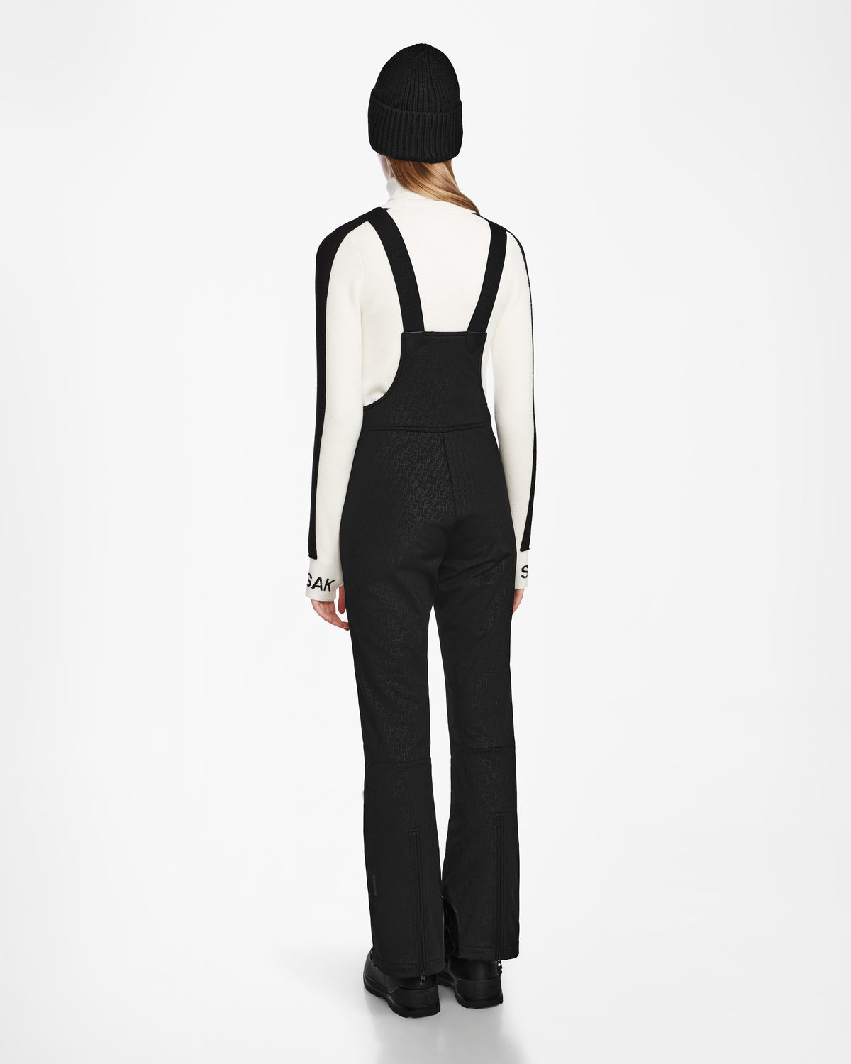 Wearing Rudsaks OKSANA - 8124527 Monogram Print Black fitted flared ski pants, a white long-sleeve top, black boots, and a black beanie, a person stands facing away against a plain white background.